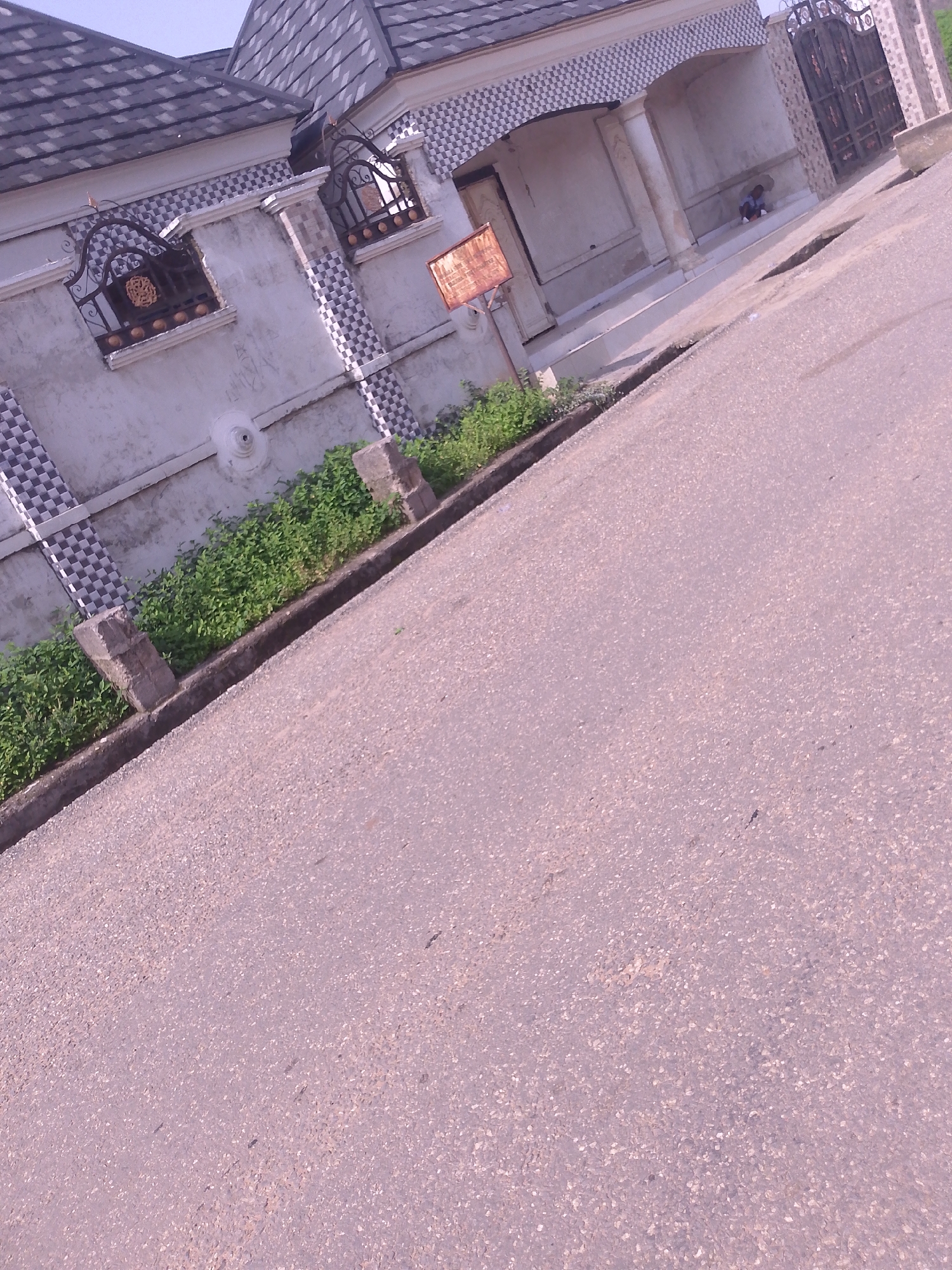 The image shows a tilted perspective of a street in Serti, Nigeria, with a building on the left and a road on the right. The building appears to be a residential house with a tiled roof, decorative window grilles, and patterned tilework. There is greenery growing along the side of the building. A person is sitting on the steps of the building's entrance. The road is paved with asphalt and shows some signs of wear. The sky is visible at the top of the image, and it is a pale blue. The overall scene suggests a sunny day in a residential area.