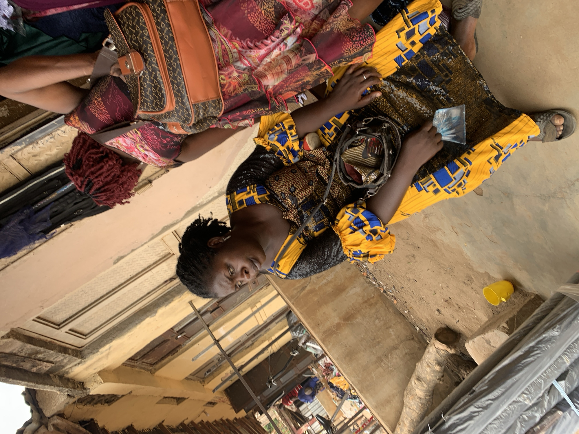 Two African women are visible outdoors. One woman sits wearing a brightly colored, patterned dress, with shades of yellow, blue, black, and brown. She holds a small package of what appears to be blades. A purse rests in her lap. She looks off in the distance. The other woman is mostly obscured. She wears a red and pink top with a bag and purse on her lap. The area appears to be a marketplace or a street market. A yellow cup and wrapped pipes are visible next to the sitting woman. The location is Kafanchan B, Nigeria. The scene is somewhat cluttered with various items and debris. The environment is gritty with dirt and visible signs of wear.