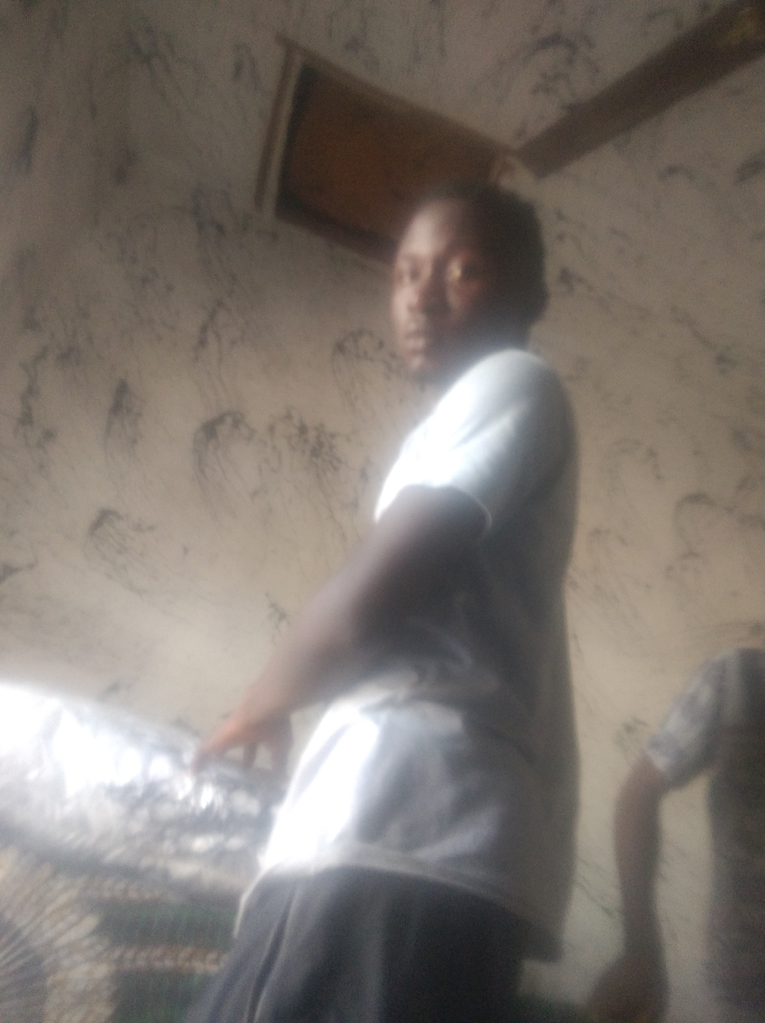 The image depicts a blurry, indoor scene in what appears to be Jalingo, Nigeria. A person, whose face is largely obscured by the blur, is visible from the chest up, wearing a light-colored t-shirt and dark pants. The individual is positioned in the foreground, facing towards the right of the frame and looking slightly over their shoulder towards the camera. The background consists of a textured wall with an abstract pattern. In the upper portion of the image, a wooden beam or plank is discernible against the wall. To the left of the main subject, there is an out-of-focus object that appears to be covered in a reflective material, possibly foil or plastic wrap, and part of a cage-like structure. Another partially visible person in a dark shirt is present in the far right of the image. The lighting is dim, contributing to the overall blurriness of the scene.