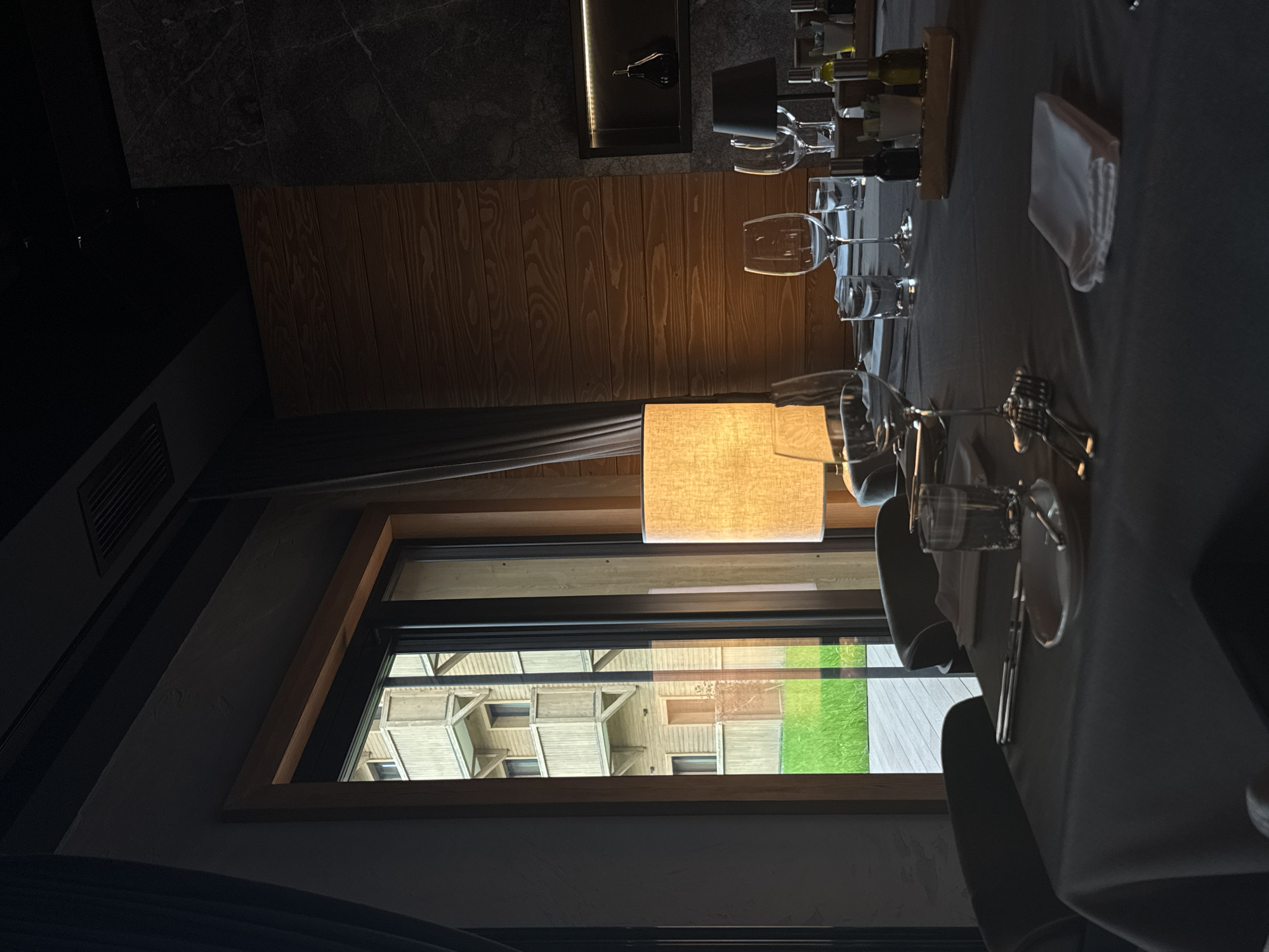The image depicts a table setting inside a restaurant or dining area in Katun Bačko brdo, Montenegro, viewed through a window. The table is set with multiple wine glasses, silverware, plates, and napkins, suggesting a formal dining arrangement. Bottles are also present, potentially wine or other beverages.
Outside the window, a structure with geometric shapes is visible, along with a patch of green grass. The window has a wooden frame. A rectangular lamp with a light-colored shade is positioned near the window, casting a warm glow.
The surrounding decor includes a wall paneling with a wood grain pattern, and darker stone or tile sections. The overall lighting in the room appears dim except for the lamp and the window.