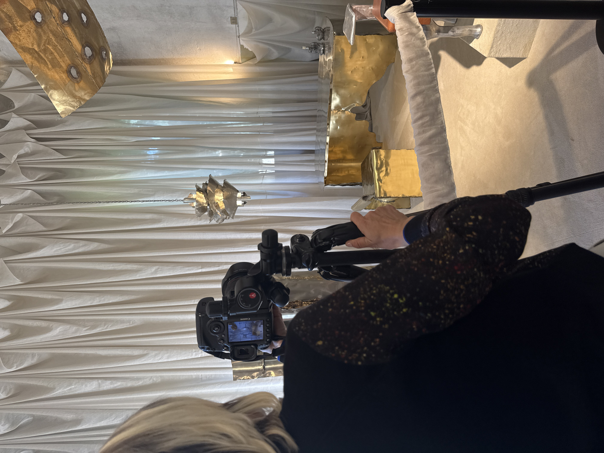 A person is using a camera on a tripod to photograph an art installation, presumably in Milan, Italy, given the prompt's request. The installation features white, draped fabric on the walls and ceiling, along with metallic, gold-colored sculptures of various shapes. One sculpture, possibly a chandelier, hangs from the ceiling. The person is wearing a dark jacket with colorful speckles. A camera screen displays a scene that closely resembles the wider shot of the art installation.