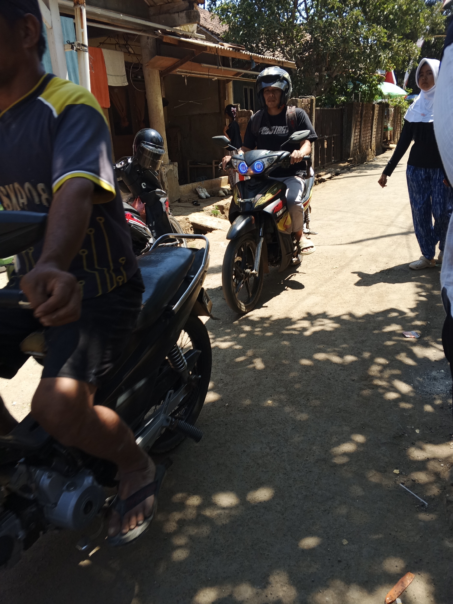 The image depicts a street scene in Sukaharja, Indonesia, on a sunny day. In the foreground, a person is seated on a motorcycle, their legs and feet visible. The motorcycle is dark-colored with red and yellow accents on the front. Further back, another person wearing a helmet is riding a motorcycle, which has blue headlights. To the right, a woman in a white hijab and dark clothing is walking away from the camera, her patterned pants visible. The background shows buildings with wooden elements and a fence. Trees cast shadows on the dusty road, suggesting a rural or semi-urban environment. The overall atmosphere is casual, with people engaged in daily activities.
