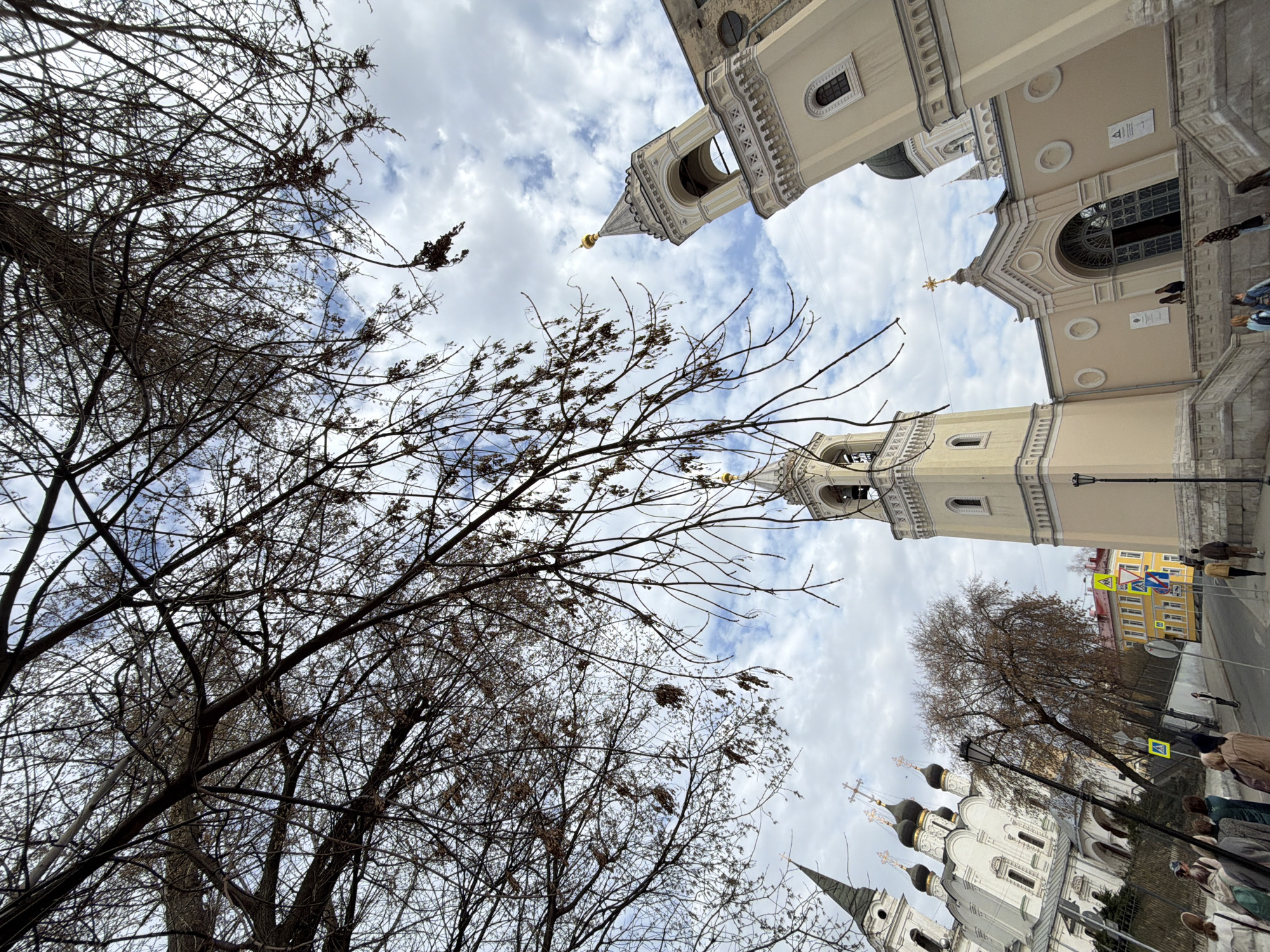 The image shows a street scene in Moscow, Russia, from a low angle, looking up at buildings and trees.
**Buildings:** There are multiple buildings, including several churches with distinct architectural styles and colors. One church has a pale yellow facade and ornate details. Another church is white with black domes and gold crosses. A tall, light-colored bell tower is also visible.
**Trees:** Bare trees with numerous branches occupy a significant portion of the left side of the image.
**People:** People are present on the sidewalk near the building, captured at ground level.
**Street:** A street and traffic signs are visible.
**Sky:** The sky is cloudy with patches of blue.