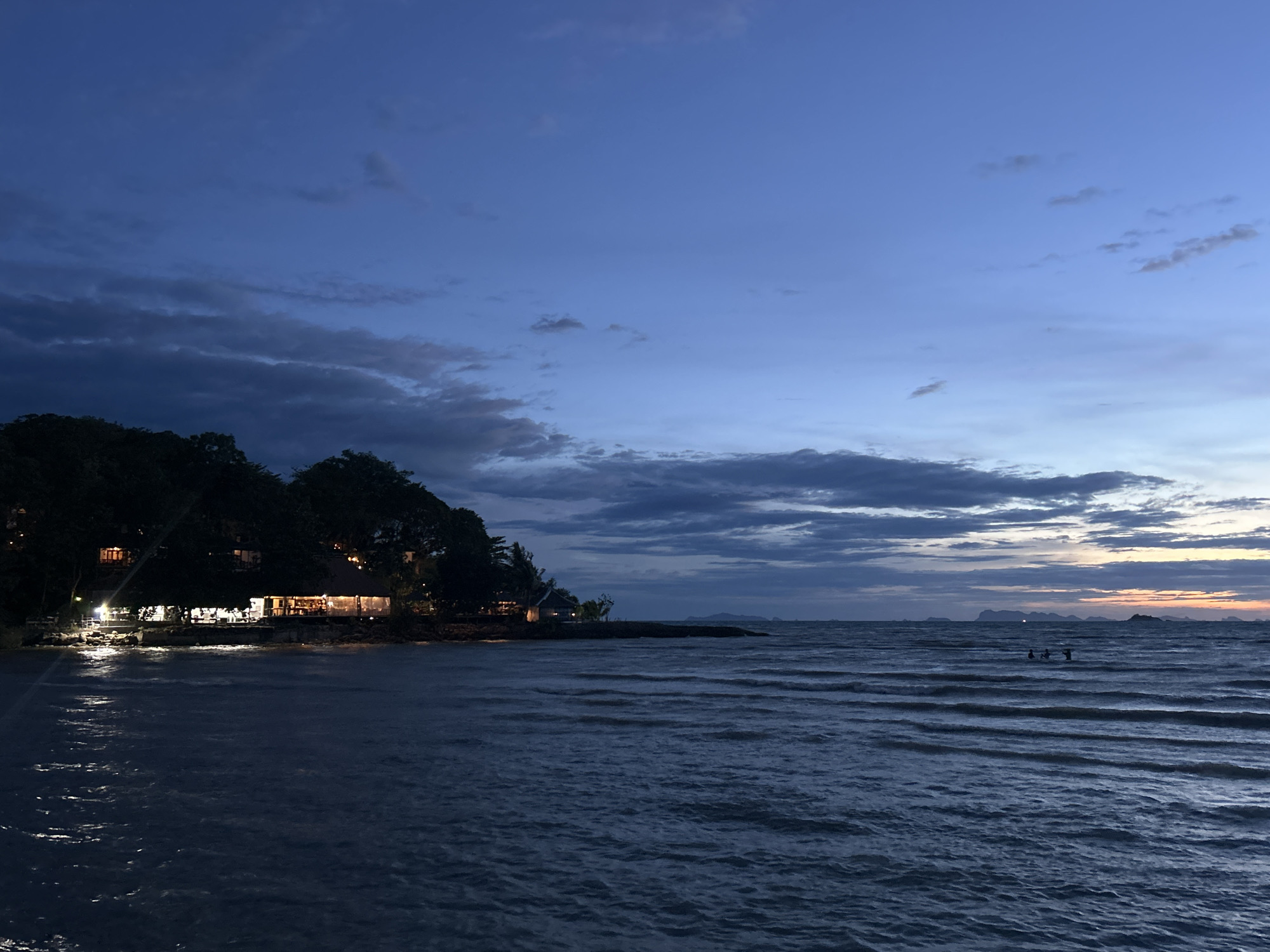The image captures a twilight scene at the beach in Baan Sri Thanu, Thailand. In the foreground, the sea displays gentle, rippling waves reflecting the ambient light. The shoreline is lined with trees, partially obscuring structures illuminated with warm lights, likely a restaurant or resort. In the mid-ground, several people appear to be wading in the water. Distant islands are silhouetted against the horizon, where the fading light of the sunset creates a soft glow. The sky transitions from a deep blue to lighter hues interspersed with cloud formations.