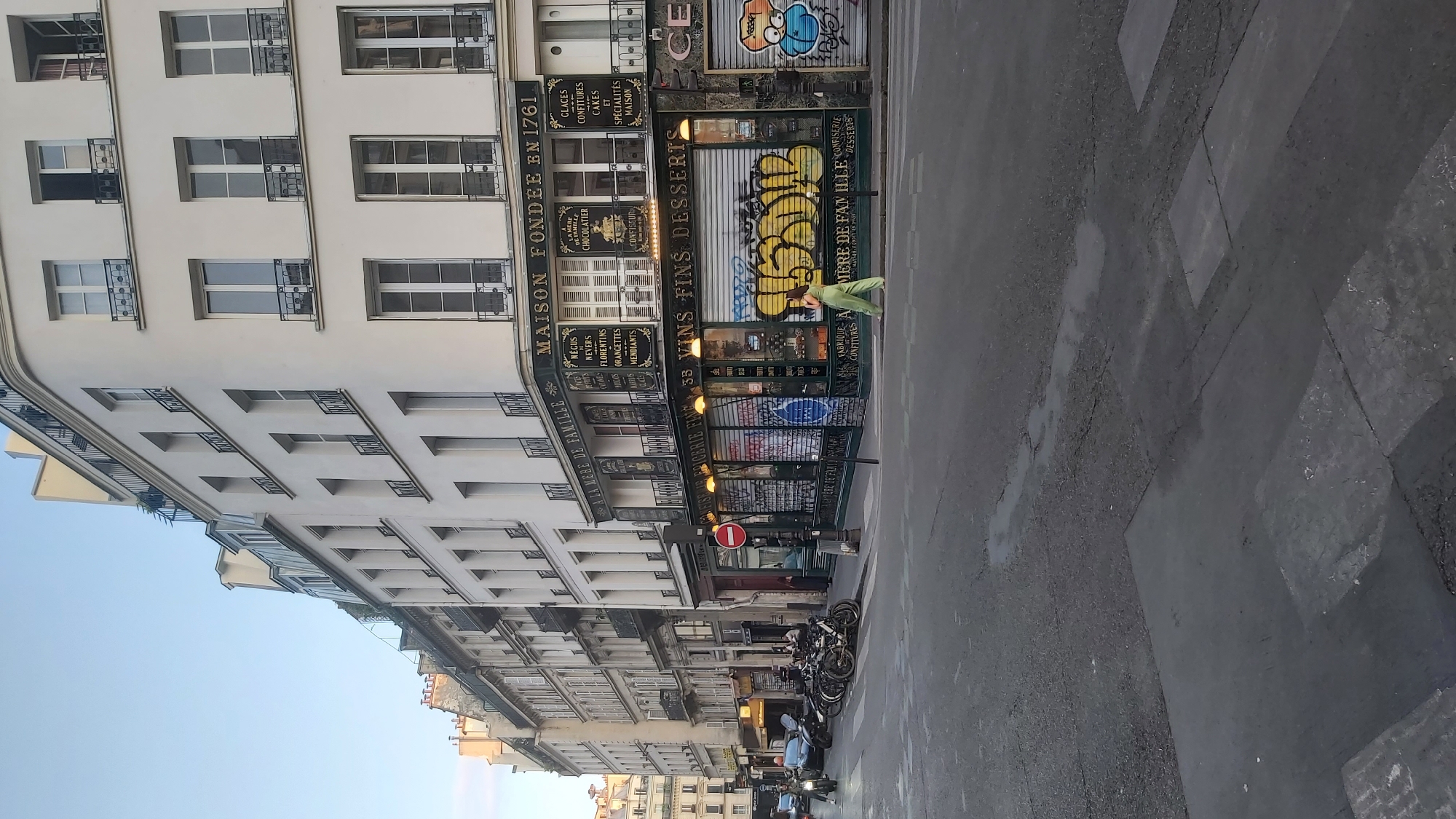 The image depicts a street scene in Paris, France, at what appears to be late afternoon or early evening, given the lighting. The perspective is from a street corner, looking down a street lined with multi-story buildings. The building on the right is a prominent feature, with a green facade featuring signs indicating it is a "Maison Fondée en 1761" and advertising various confectioneries and desserts. The ground floor of this building has roll-down shutters adorned with graffiti. A woman in a bright green outfit is walking across the street in front of this building.
Further down the street, to the left, are more buildings with balconies and windows typical of Parisian architecture. Several parked motorcycles and bicycles are visible on the sidewalk. Traffic signals and street signs are present. The street itself is paved with asphalt and shows signs of wear, with some puddles. The sky is clear and blue. The overall impression is of a typical Parisian street with a blend of historical architecture and modern elements like graffiti.