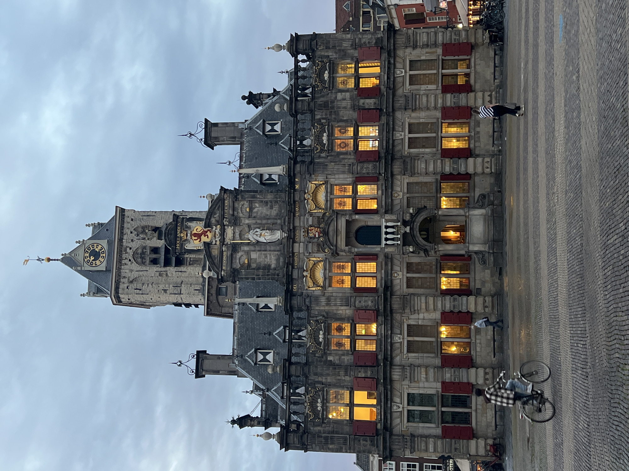 The image shows the Market Square in Delft, Netherlands, in the late afternoon or early evening. The focal point is the historic Delft City Hall, a grand Renaissance building with a prominent clock tower. The facade is made of dark stone, intricately carved, with many windows. Warm light emanates from some of the windows, suggesting occupancy. Red shutters adorn many of the windows, adding a splash of color to the stone.
In the foreground, the cobblestone square is visible. Two people are present. On the left, a person on a bicycle rides from left to right across the frame, wearing a plaid shirt and jeans. On the right, a person walks away from the camera towards the building, wearing a striped shirt and dark pants, carrying a bag. The sky is overcast, with a soft, diffused light. The overall atmosphere is calm and somewhat subdued, characteristic of the twilight hours in a historic European city.