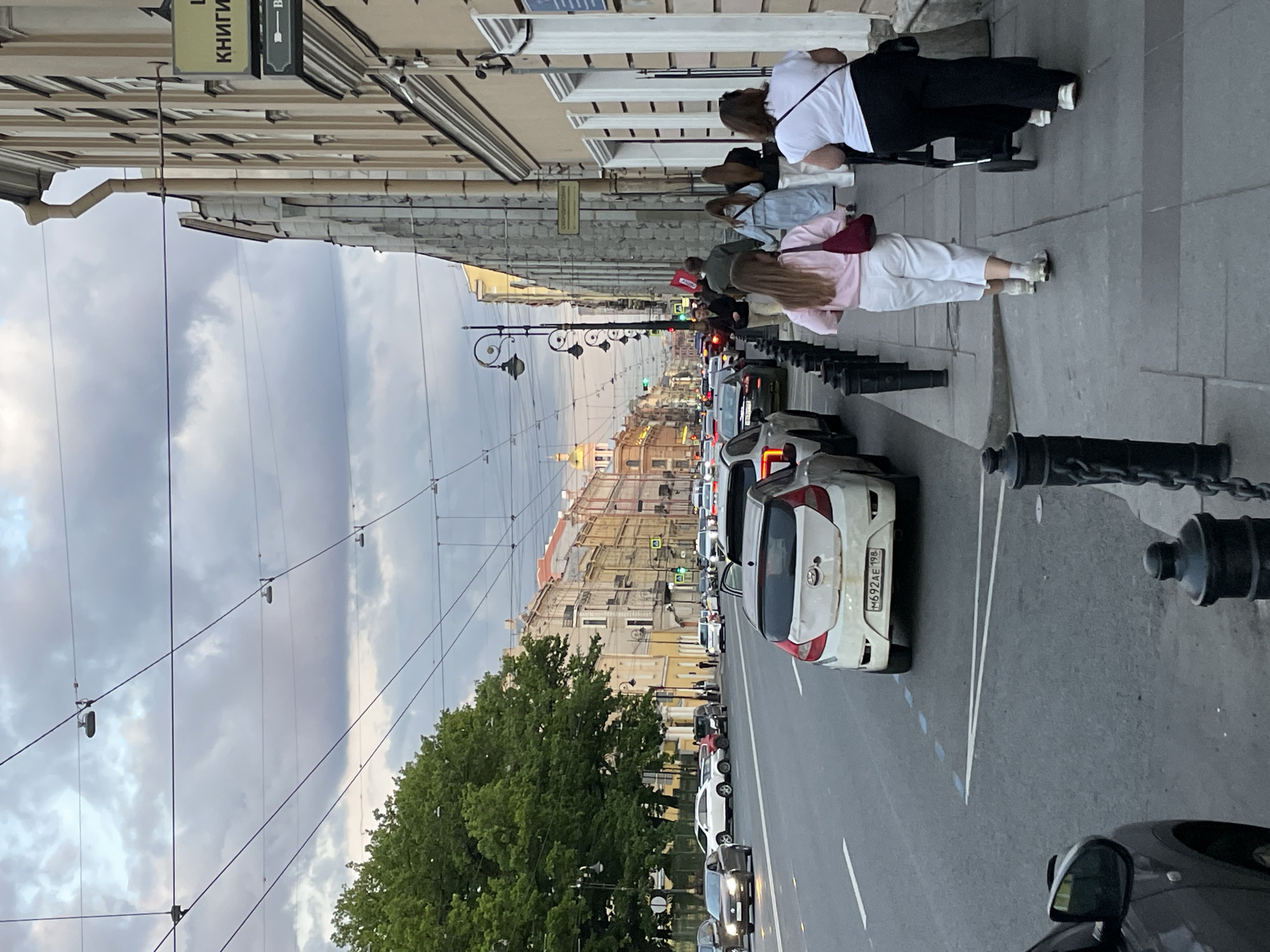 This image captures a street scene in Saint Petersburg, Russia, likely during the late afternoon or early evening under an overcast sky. The photo is rotated 90 degrees clockwise.
On the left side of the frame, historical buildings with detailed facades line the street, featuring windows with awnings. A sign hanging from one building clearly displays "КНИГИ" (KNIGI), meaning "BOOKS" in Russian, next to another sign with an arrow pointing right. A wide sidewalk runs along these buildings, where several people are walking. One person in a white t-shirt and black pants is visible at the top, while further down, another person in a pink top and white pants with a red fanny pack is walking. Black ornate streetlights are periodically placed along the sidewalk.
The right side of the image shows a multi-lane road with moderate traffic. A white Toyota Yaris hatchback is prominently featured, displaying a Russian license plate "H692AE 198," with "198" confirming the Saint Petersburg region. Other cars, including a dark vehicle in the immediate foreground, are visible on the road, suggesting typical urban activity. Black bollards with chains line the edge of the sidewalk, separating it from the street.
Overhead, numerous wires crisscross the sky, characteristic of cities with trolleybuses or trams. In the distance, the street curves, and more buildings, some with domes, are visible under the cloudy sky, alongside green trees. The overall scene depicts a bustling urban environment.