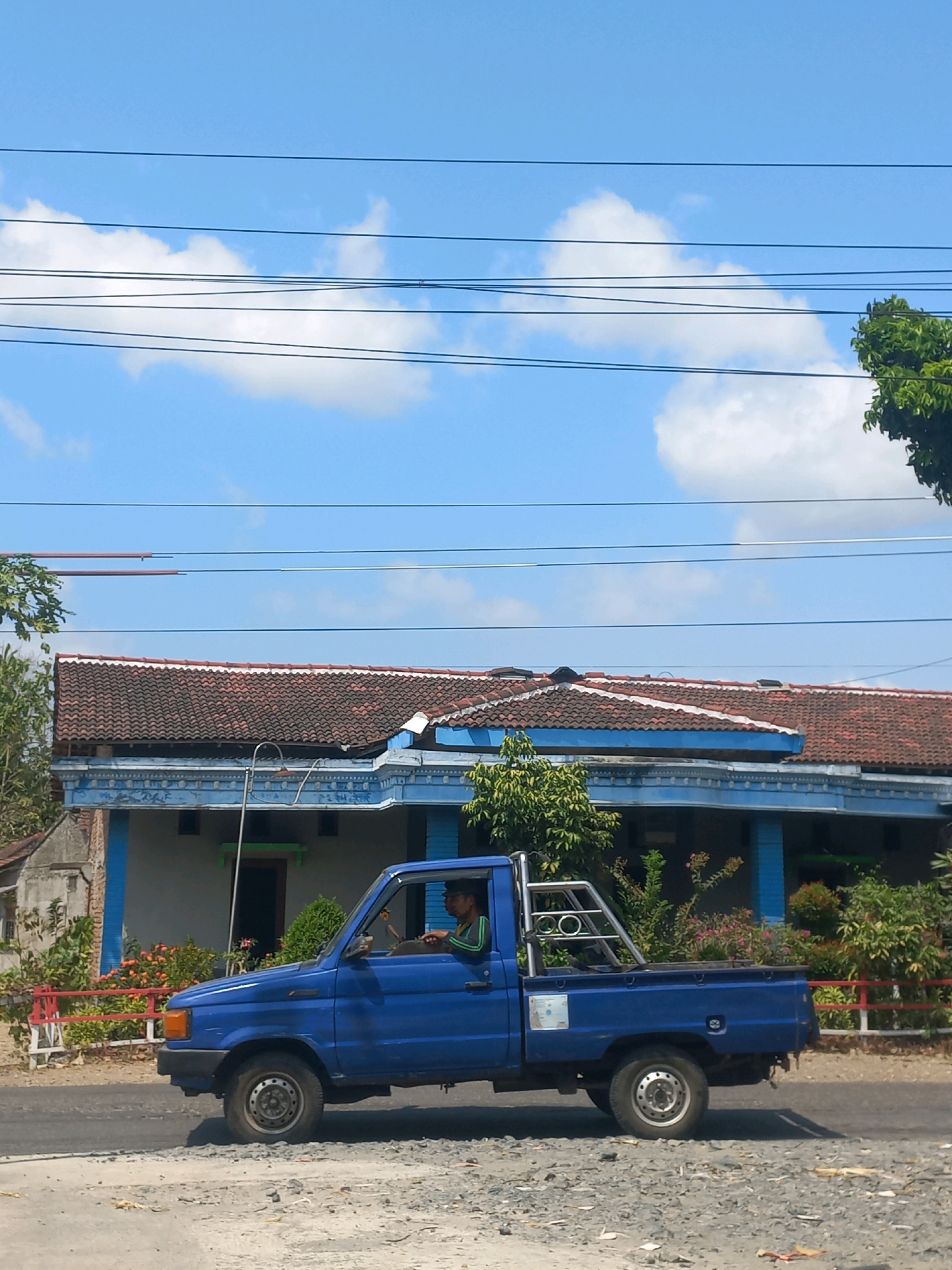 A blue pickup truck with a driver is shown in profile, moving from left to right on a paved road in Baturetno, Indonesia. The truck has a silver roll bar in the bed. The driver, a person with dark hair and wearing a green shirt with dark stripes on the sleeves, is looking forward. In the background, a house with a tiled roof and blue trim is visible. The house is surrounded by greenery, including trees and flowering bushes. Several power lines are strung across the bright blue sky, which is dotted with white clouds. The ground in the foreground is a mix of pavement and gravel.