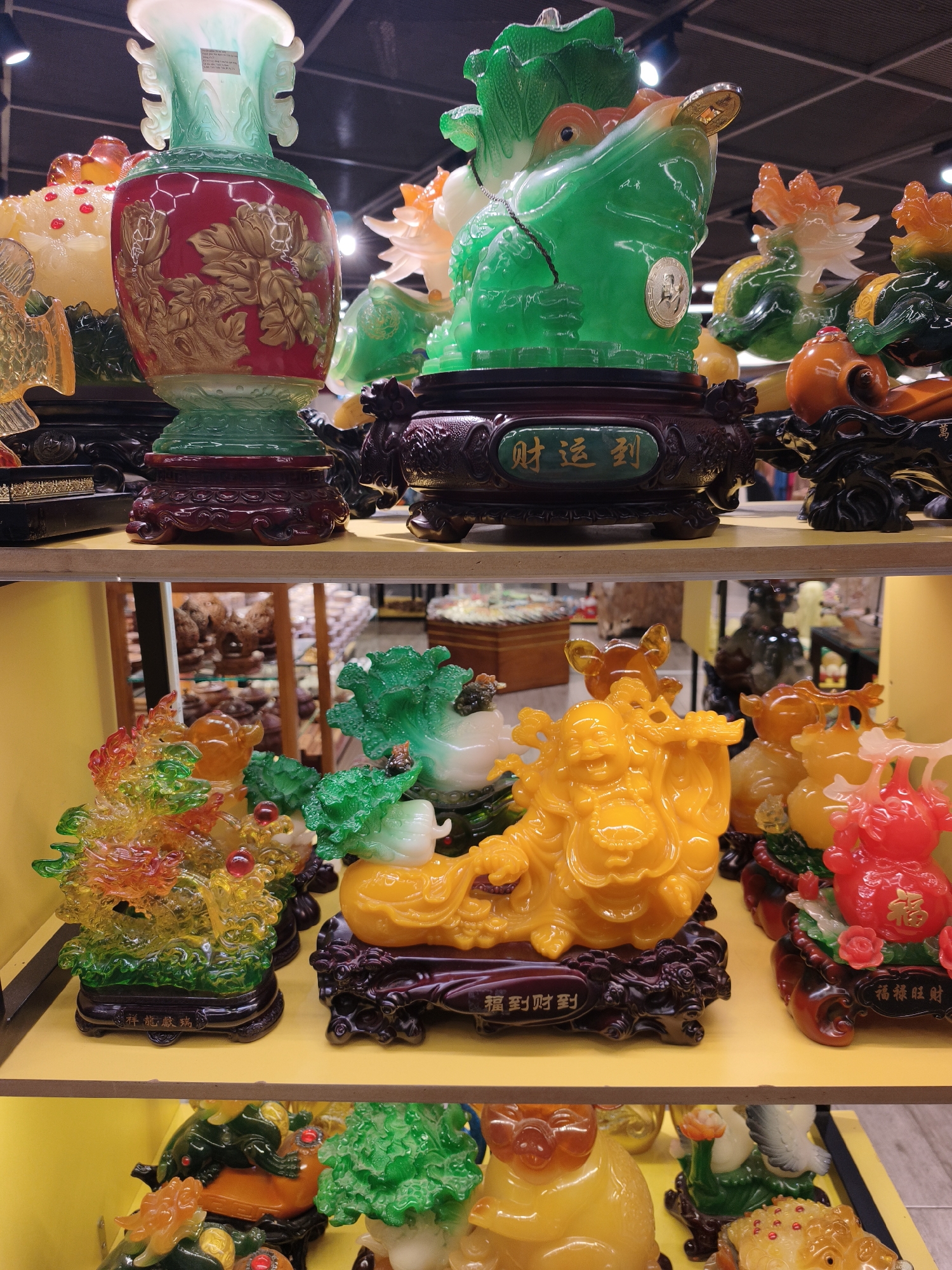 The image displays a close-up, eye-level, indoor shot of shelves laden with colorful, decorative figurines, likely from a shop in Nha Trang, Vietnam. The objects are primarily made of resin or a similar material, with a glossy finish.
The top shelf features a large, translucent jade-green and red vase with intricate gold floral motifs. To its right is a prominent, oversized green frog figurine with a coin in its mouth, sitting on a dark wooden base with Chinese characters. Behind it, other brightly colored translucent animal figurines, possibly dragons or phoenixes, are visible.
The middle shelf showcases more elaborate resin sculptures. On the left, a cascading arrangement of red, orange, and green translucent elements, resembling coral or flames, sits on a dark base with Chinese characters. To its right, a smiling, plump yellow figurine, resembling the Laughing Buddha, is depicted holding a pouch, also on a dark, ornate base with Chinese characters. Further to the right, a bright red figurine, possibly a pig or a similar auspicious animal, adorned with roses, rests on a decorative base.
The bottom shelf, partially visible, shows more of these colorful figurines, including a green frog and what appears to be a larger version of the yellow Laughing Buddha-like figure. The shelves are made of a light-colored wood, and the background suggests a retail environment with other displays or products blurred out. The lighting is bright, illuminating the vibrant colors of the figurines. There are no people visible in the frame.