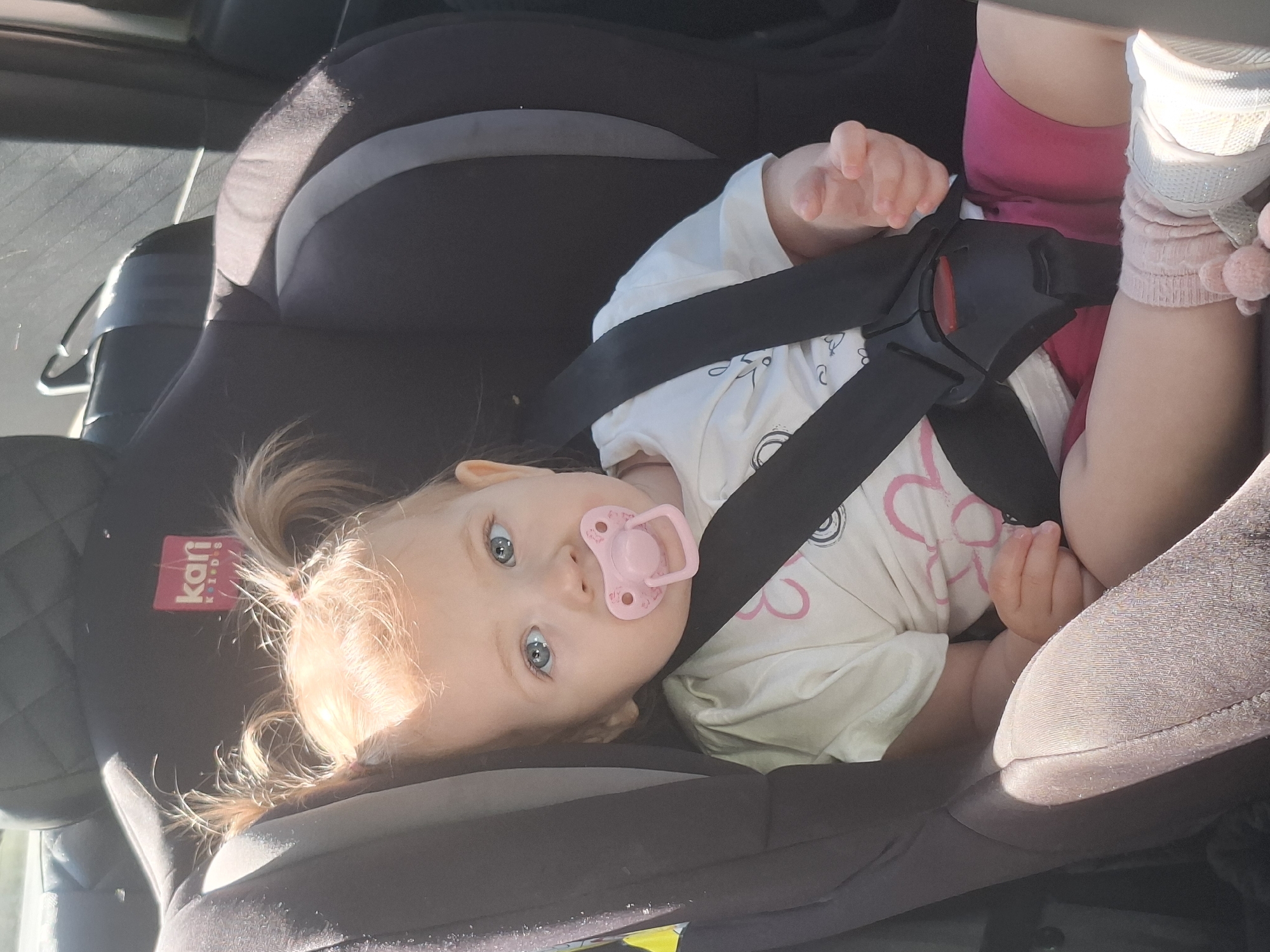 A baby with blonde hair in pigtails is secured in a black and grey car seat with black safety straps. The baby is wearing a white shirt with pink and black drawings, pink shorts, white shoes, and pink socks with pom-poms. The baby has a pink pacifier in its mouth. The brand "Kari Kids" is visible on the car seat. The photo appears to have been taken inside a car, with part of the interior visible. The location is Chaikovsky, Russia, based on the information on the car seat.