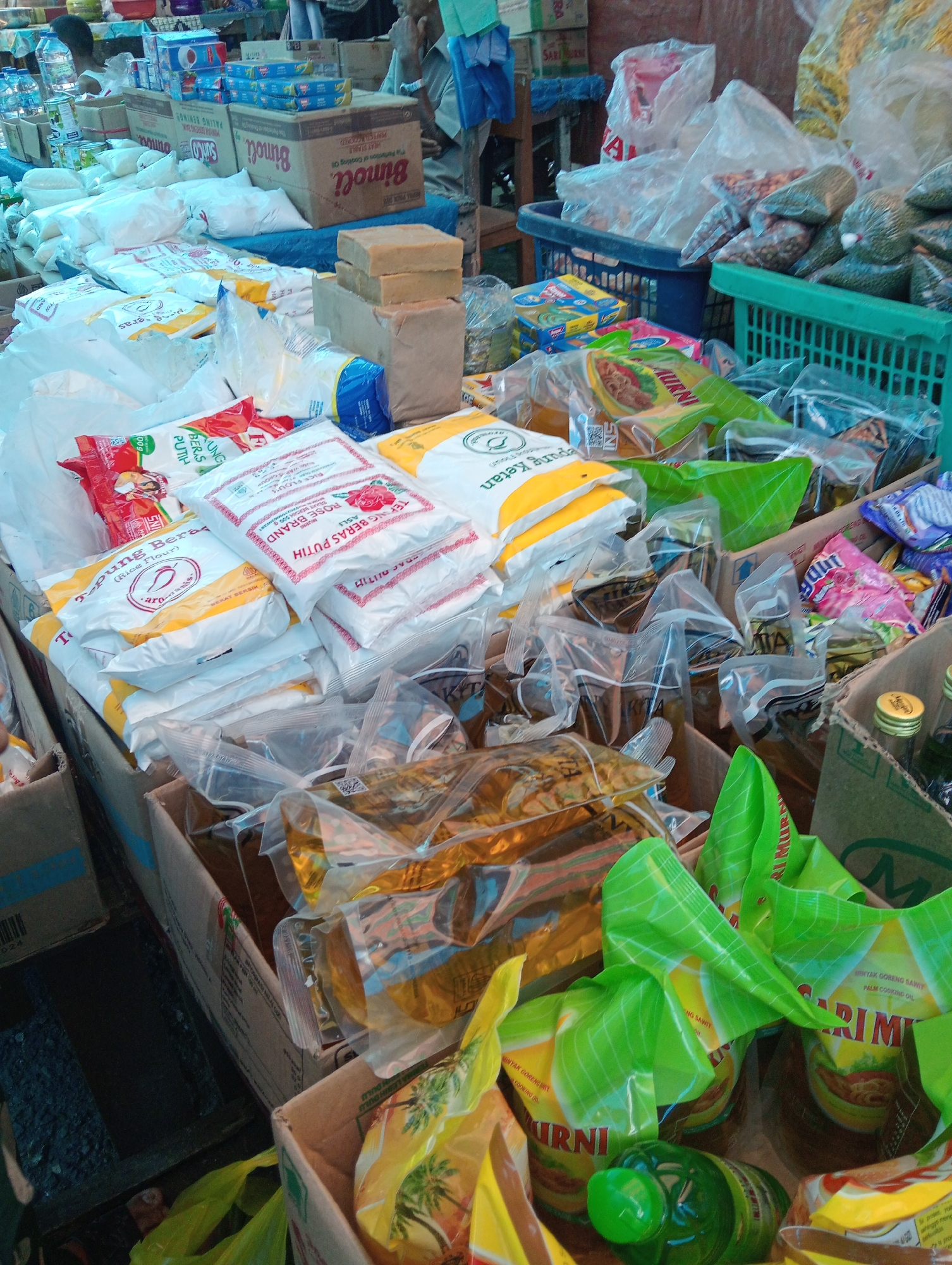 The image depicts a street-level view of a market stall in Pariaman, Indonesia, filled with an assortment of packaged goods. Dominating the foreground and midground are stacks of white and yellow bagged products, many with red rose branding and text indicating "Tepung Beras PUTIH" (White Rice Flour) and "Rose BRAND". Interspersed are various other bagged items, some in clear plastic with visible contents, and some with colorful branding, including what appears to be cooking oil. To the right, green plastic baskets contain more bagged items, and in the background, several cardboard boxes are visible, including one with "Binoli" branding. The overall scene suggests a bustling market environment, likely selling food staples and household items. No people are clearly visible or engaged in any specific actions within the frame. The lighting suggests it is daytime, with natural light illuminating the scene.