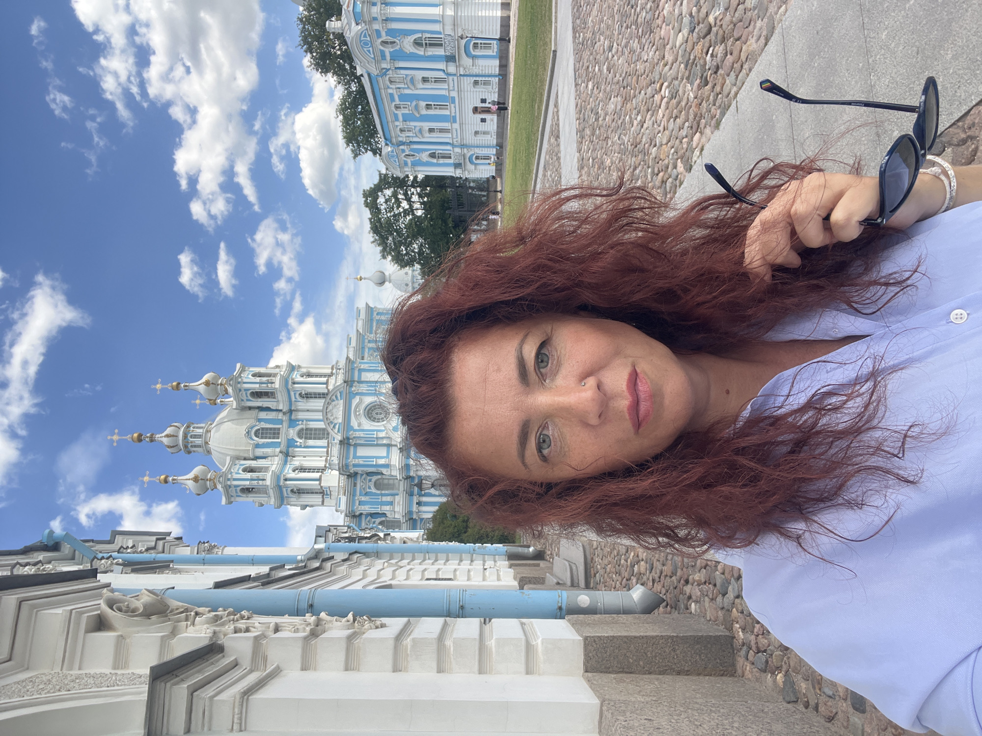 A woman with long, reddish-brown, curly hair is taking a selfie in front of the Smolny Cathedral in St. Petersburg, Russia. She is wearing a light blue collared shirt and holding a pair of black sunglasses in her right hand. The Smolny Cathedral, a prominent blue and white Baroque building with multiple domes, is visible in the background, along with a cloudy blue sky. The foreground is a cobblestone pathway.