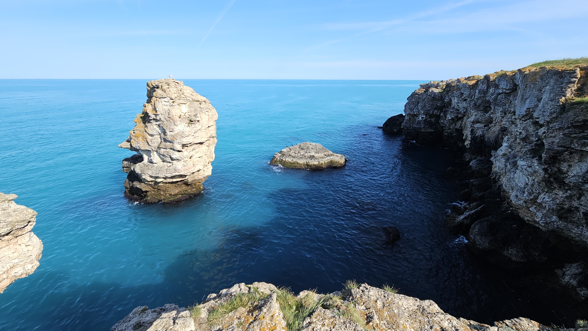 A high-angle scenic view captures a coastal landscape. The scene includes a tall, weathered rock formation standing in turquoise waters near the center-left. Another smaller rock is near it. To the right, rugged cliffs rise above the darker blue water. The foreground is bordered by rocky terrain. The sky is clear and blue.