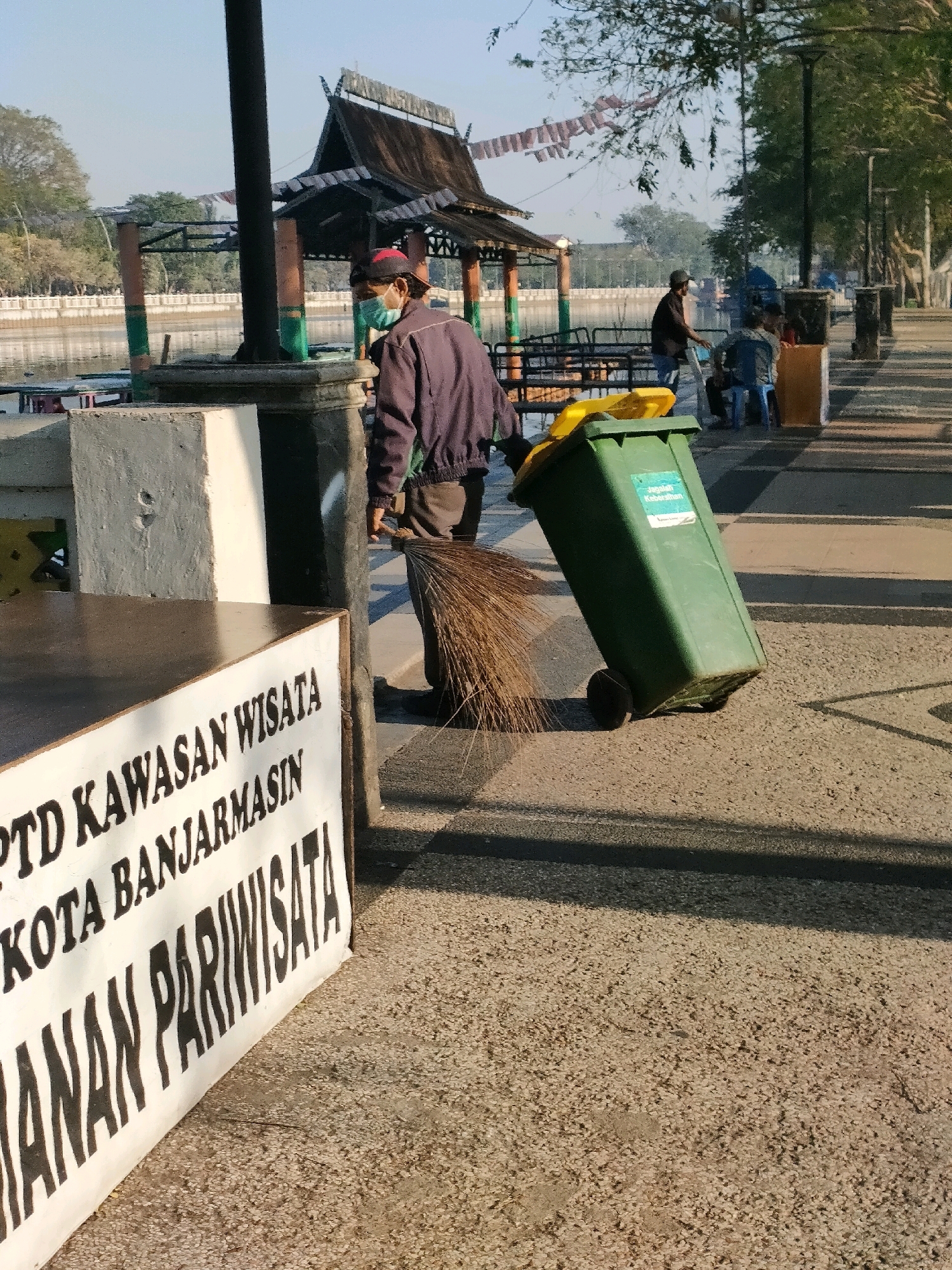 The image depicts a sunny outdoor scene in Banjarmasin, Indonesia, likely in a park or waterfront area. A sanitation worker, wearing a face mask and a red cap, is pushing a large green wheeled trash bin with a yellow lid. He is also holding a broom. In the background, there is a body of water, a covered pavilion with traditional architecture, trees, and other people who appear to be relaxing. A white sign with black text is prominently placed in the foreground, partially obscuring the trash bin. The text on the sign, in Indonesian, reads "PTD KAWASAN WISATA KOTA BANJARMASIN" and "ANAN PARIWISATA", indicating a tourist area in Banjarmasin. The ground is paved with a textured surface, and shadows are cast by the sun, suggesting it is either morning or late afternoon.