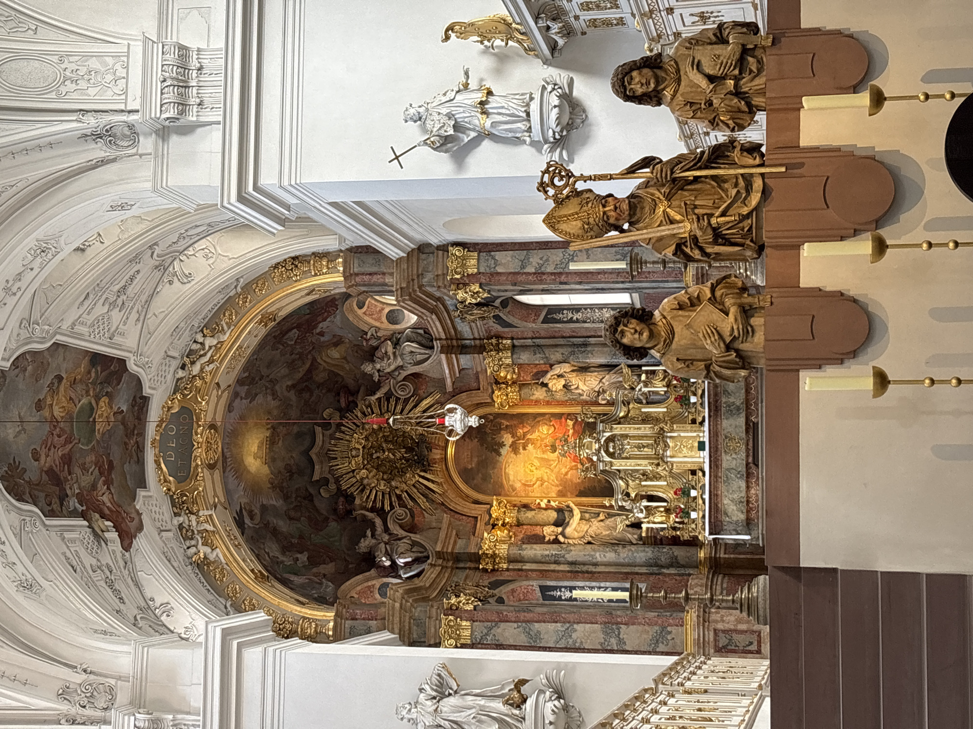 This image shows the interior of the Würzburg Residence Chapel, a Baroque chapel located in Würzburg, Germany. The chapel is elaborately decorated with frescoes, sculptures, and gilded ornaments. In the foreground, there are three wooden sculptures of saints, each holding a book. The central sculpture depicts a bishop wearing a mitre and holding a crosier. Behind the sculptures is the main altar, which is adorned with intricate gold carvings and religious paintings. The altar is illuminated by a bright light, creating a dramatic effect. The ceiling is painted with a fresco depicting biblical scenes, and the walls are decorated with white stucco and gold trim. The overall impression is one of grandeur and opulence, typical of Baroque religious architecture.