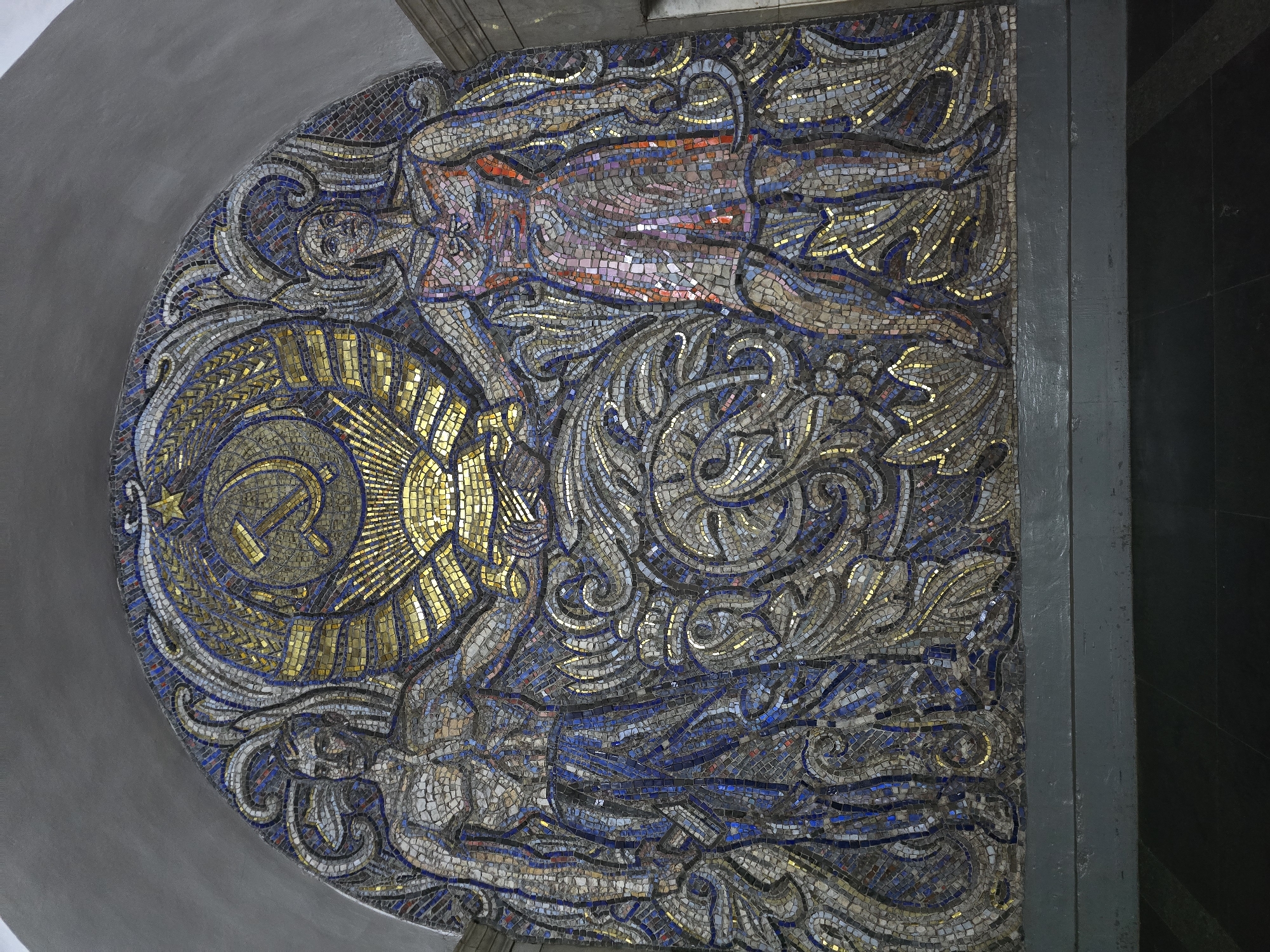 This is a photograph of a mosaic art piece located in Moscow, Russia. The mosaic is an arched shape, likely filling the space above a doorway or entryway. The primary colors are gold, blue, silver, and shades of grey, with small sections of orange and red. The mosaic depicts two human figures surrounded by swirling, decorative patterns.
On the left side of the arch is a gold-colored symbol of the hammer and sickle, a symbol of communism. Rays emanate from this symbol, giving it a sun-like appearance. Above the symbol is a figure rendered in shades of orange and red with blue and grey detailing. Below the symbol is another figure rendered in silver, blue, and grey tones. The swirling patterns surround the figures and the central symbol, creating a sense of movement and depth. The mosaic is composed of small, individual tiles. The mosaic is set into a wall, which is painted grey above and to the right with a black or dark grey vertical support beam on the right side.