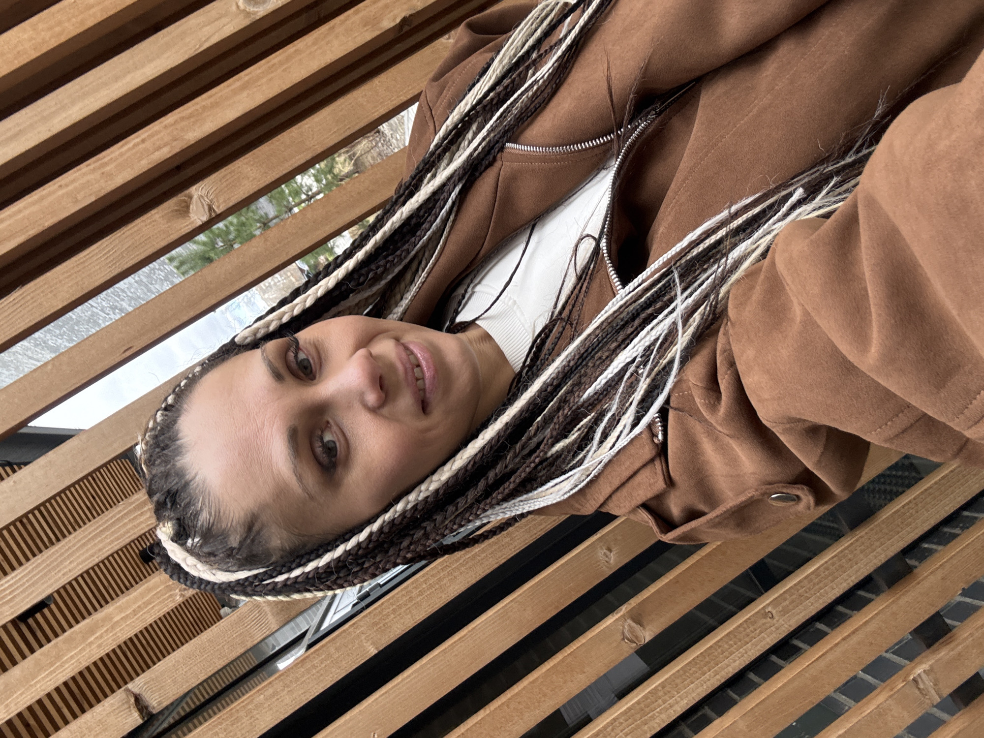 A medium shot depicts a woman with tan skin, wearing brown and white braided hair, and a brown suede jacket with a white undershirt. She is lying on a horizontal wooden latticework, potentially a fence or decorative feature. The background shows blurred glimpses of trees and a window, suggesting an outdoor or semi-outdoor location in an urban area. The city is Unknown, Unknown.