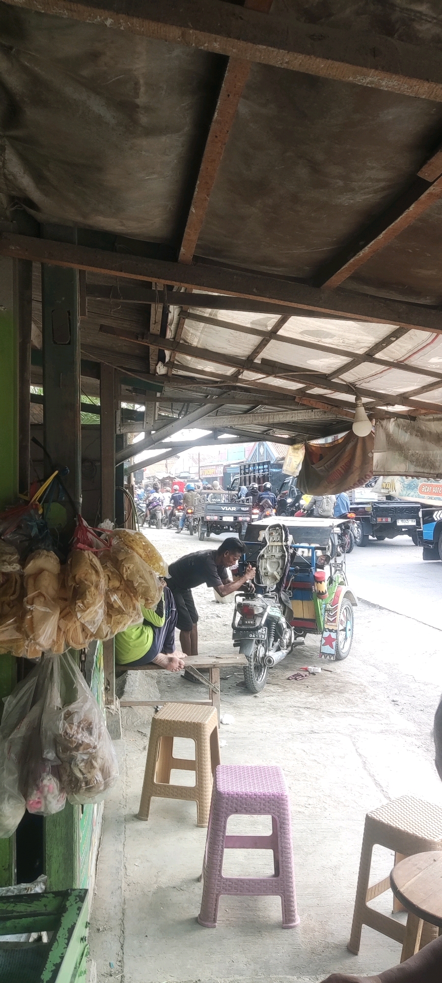 This image depicts a roadside scene in Gebang, Indonesia, under a makeshift shelter. A man in a dark t-shirt is leaning over a three-wheeled motorcycle, appearing to be working on it. Next to him, a person wearing a green shirt is seated on a wooden bench. The shelter's roof is constructed with corrugated metal sheets supported by wooden beams. In the foreground, two plastic stools, one beige and one pink, are positioned on the concrete ground. Bags of what appear to be snacks or food items are hanging from a green structure on the left side of the frame. The background shows a street with various vehicles, including motorcycles and trucks, indicating a busy commercial or transportation area. The lighting suggests it is daytime.