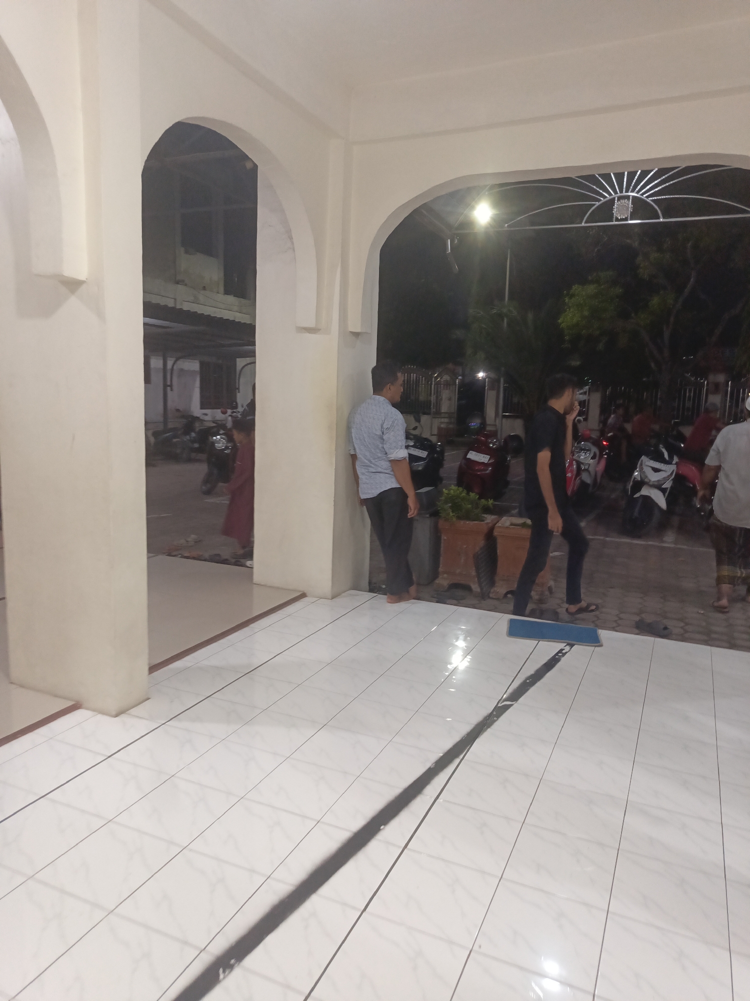 The image depicts an outdoor scene at night, likely near a mosque or a community gathering space, given the presence of motorcycles and architectural elements that suggest a religious or communal building. Several individuals are visible. On the left, a person in a maroon outfit stands near an arched doorway, looking into an interior space where several motorcycles are parked. To the right of this doorway, a man in a light blue shirt and dark pants leans against a wall. Further to the right, another man in a black shirt and dark pants is walking, with a mop and bucket visible on the tiled floor in front of him. Behind him, more motorcycles are parked. The ground is tiled, with dark grout lines, and a long shadow is cast across it, indicating artificial lighting. The overall atmosphere appears calm, with the subjects seemingly engaged in ordinary activities like waiting, walking, or cleaning. The lighting suggests it is nighttime, and the presence of lush trees in the background hints at a warm climate. The location is identified as Lhokseumawe, Indonesia. No text is visible in the image.