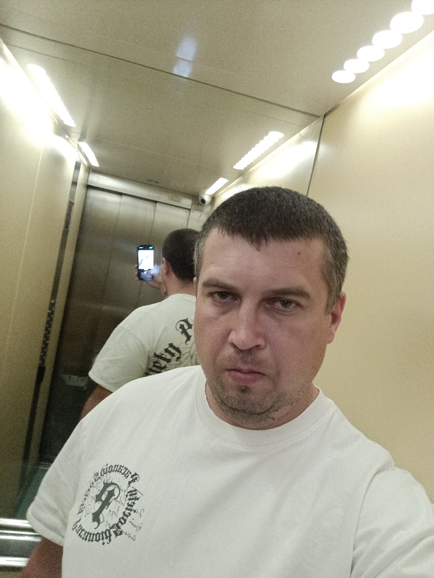 A man in a white t-shirt is taking a selfie in an elevator. The man has short, dark hair and a light stubble. He is looking directly at the camera with a neutral expression. Behind him, a reflection in the elevator doors shows another man, also in a white t-shirt, holding a smartphone and taking a picture. The elevator interior is illuminated by bright fluorescent lights on the ceiling and walls. The walls are a pale yellow, and there are metal fixtures visible, including elevator doors and what appears to be a security camera. The t-shirt in the foreground has a graphic design on the left chest with text in a gothic font, though it is partially obscured. The reflection of the other man's t-shirt also shows a similar graphic with text, which appears to read "Society P" and other unreadable characters. The scene is likely taking place indoors, possibly in a hotel or public building, given the elevator setting. There are no clear indicators of the time of day or weather conditions. The overall mood is casual and everyday.