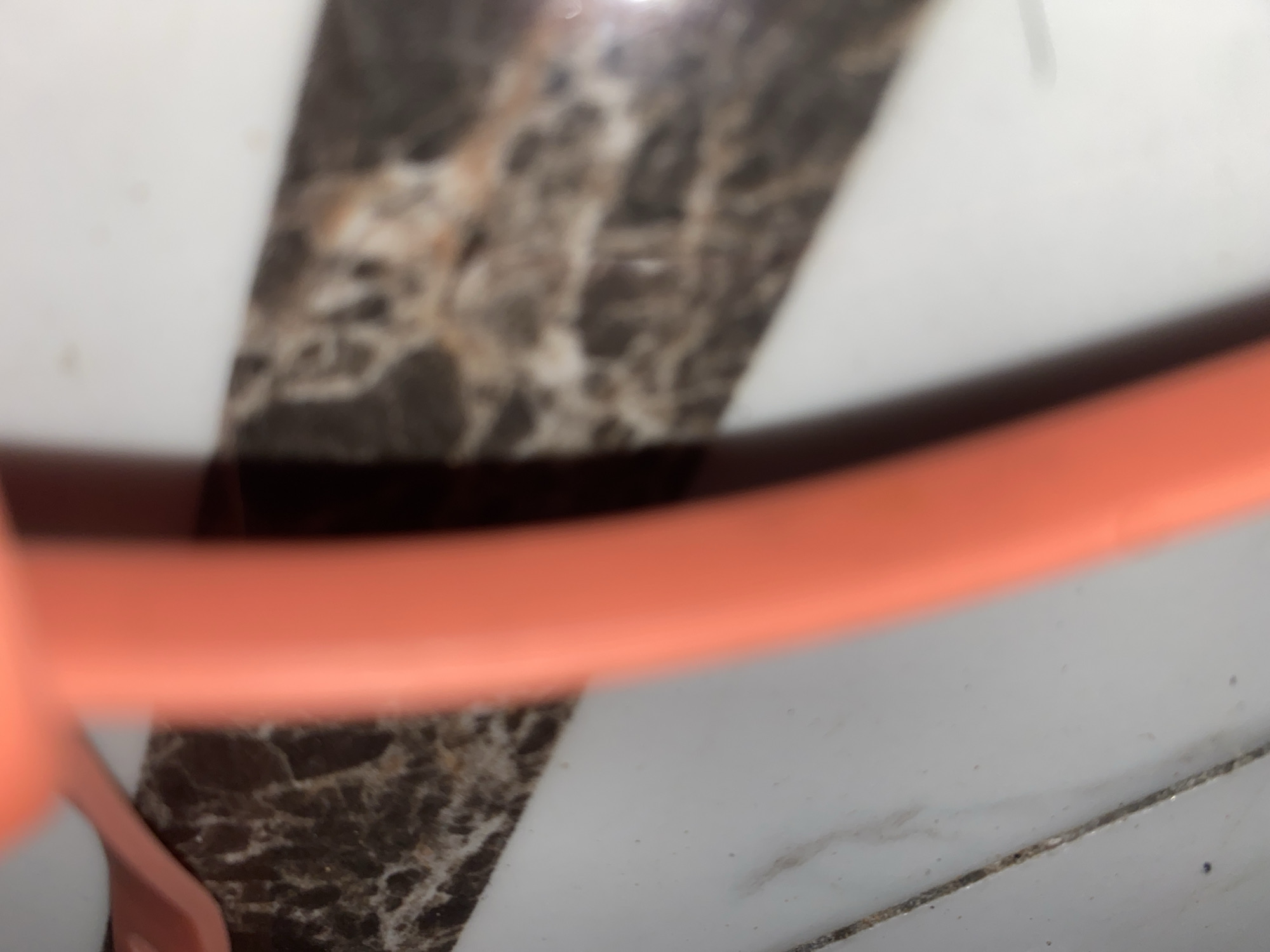 The image is an extreme close-up, out-of-focus shot of an orange object, likely a piece of plastic, lying on a tiled surface. The tiles appear to be a light color with dark, marbled veins. The orange object is curved and takes up a significant portion of the frame, obscuring much of the background. No people or specific landmarks are visible. The scene is generic and could be anywhere, including Jauro Yinu, Nigeria, as the surface appears to be a common indoor or outdoor flooring material.