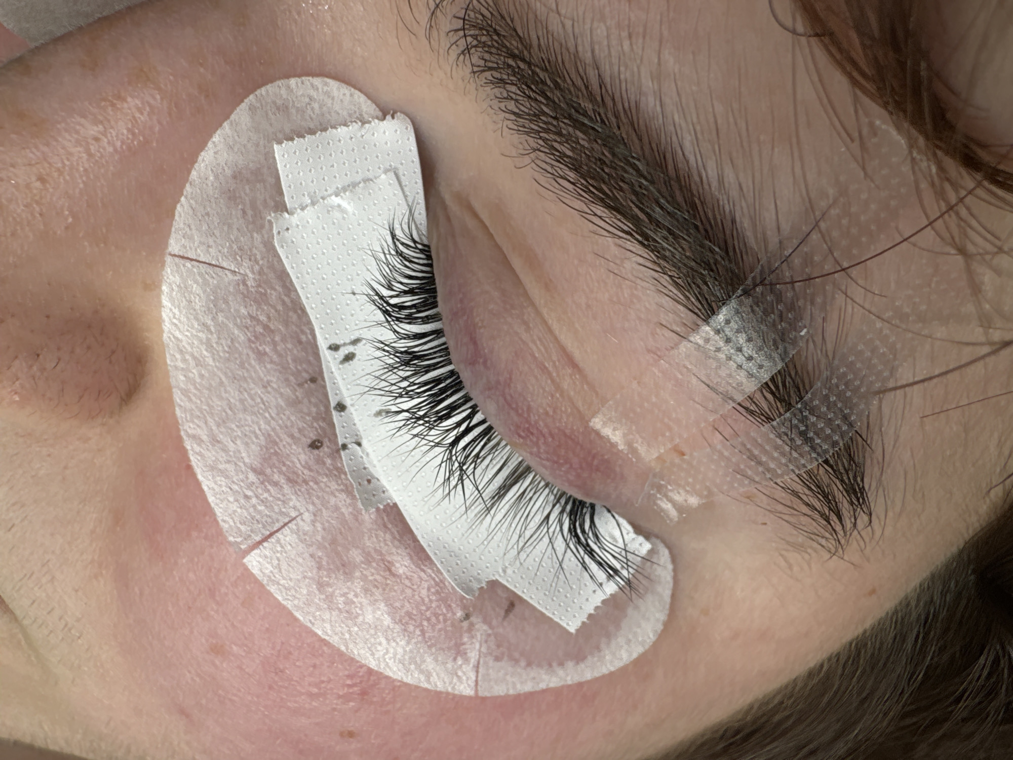 This is a close-up, forensic photograph of a person's eye and eyebrow area, possibly taken in a beauty salon. The subject's eye is closed, and individual false eyelashes have been applied to the natural lashes. Protective under-eye pads, secured with medical tape, are visible on the skin below the lower lash line. The skin appears slightly reddened. The eyebrow above the eye is dark and groomed. The image captures a detailed view of cosmetic procedures being performed. No specific location in Moscow, Russia, can be determined from the visual information provided.