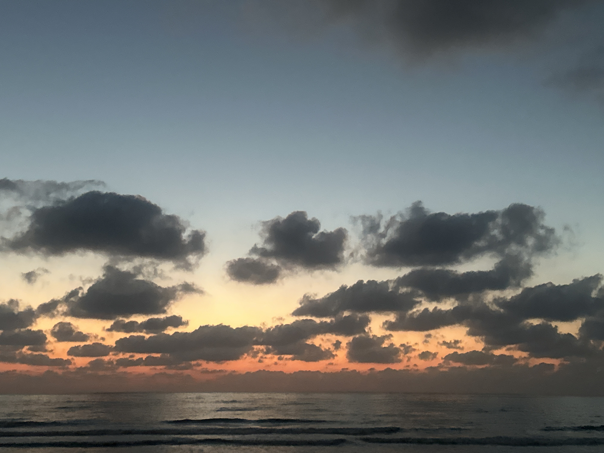 This media file captures a serene seascape at dusk. The scene is dominated by a vast expanse of ocean meeting a sky filled with dramatic clouds. The time of day is evident from the soft, warm hues of orange and yellow painting the horizon, transitioning into cooler blues and grays higher in the sky. These colors suggest the sun is either rising or setting, creating a picturesque twilight atmosphere.
The sky is populated by various types of clouds. Some are dark and ominous, casting shadows and adding depth to the composition, while others are lighter, catching the ambient light and appearing almost wispy. The clouds are densely packed in the middle and lower sections of the sky, gradually thinning out towards the top.
The ocean in the foreground appears calm, with gentle waves creating subtle ripples and horizontal lines across the water's surface. The water reflects the muted colors of the sky, adding to the tranquil and somewhat melancholic mood of the scene. There are no visible people or man-made objects, focusing entirely on the natural elements of the sky and sea.
The location context of Haifa, Israel, brings a potential cultural association to mind, but the image itself is a universal depiction of coastal beauty during transitional light. The overall emotion evoked is one of peace, contemplation, and the sublime beauty of nature. There is no visible text within the image.
