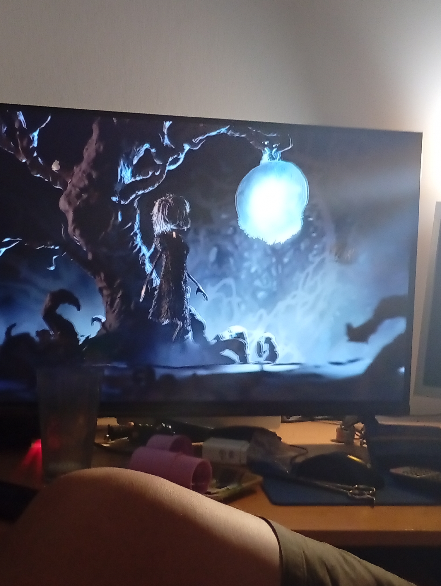 A screen displaying a scene of a tree, a humanoid figure, and a bright orb is visible. Below the screen is a desk cluttered with objects including a drinking glass, pink hair curlers, a mouse, a mousepad, and scissors. A person's leg is in the foreground. The location in Buenos Aires, Argentina is not discernible from the image.