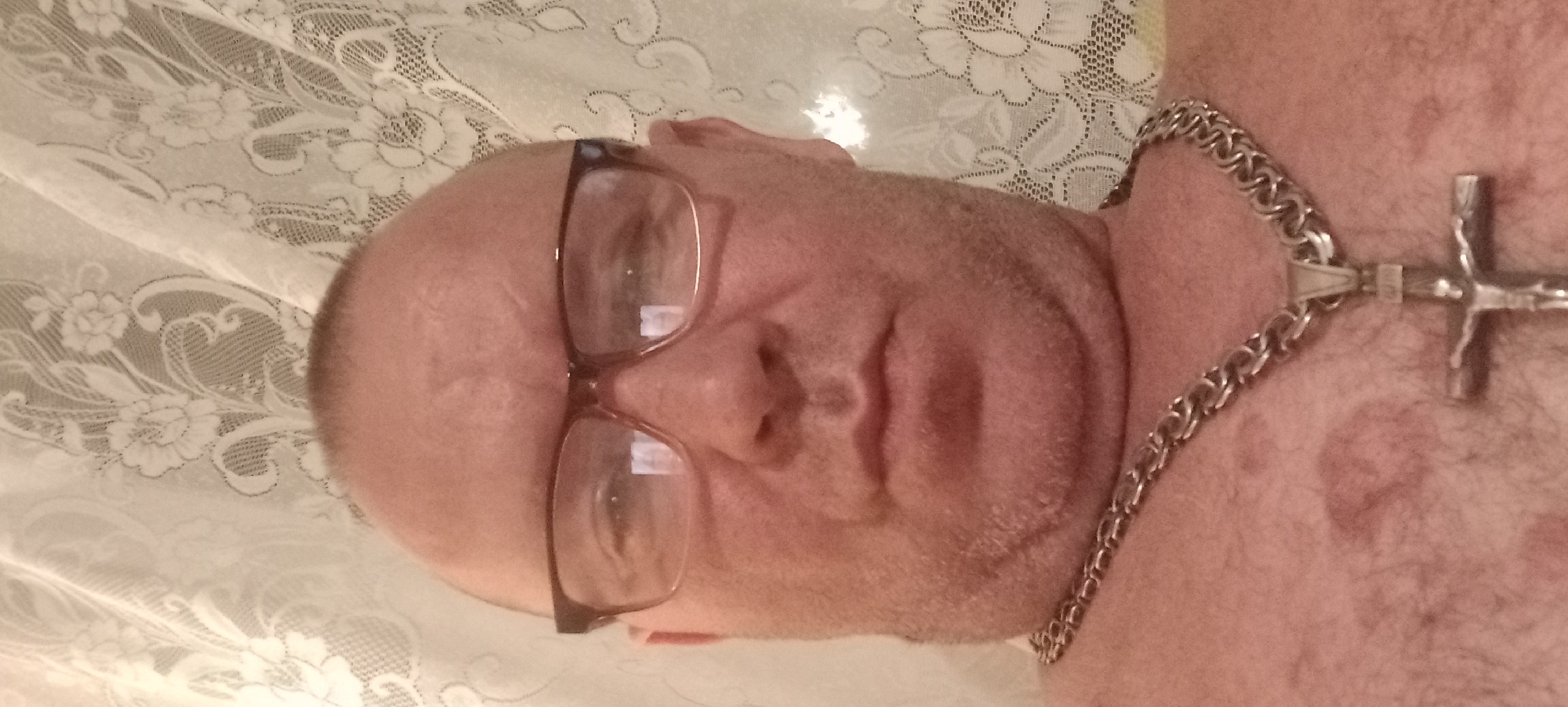 The image is a close-up selfie of a bald man wearing glasses and a silver chain with a crucifix. The man has a fair complexion and is looking directly at the camera. He has stubble on his face and appears to have some marks on his chest. He is wearing a necklace with a large, silver crucifix. The background is a white lace curtain with a floral pattern. The location is stated as the city of Kryvyi Rih, Ukraine.