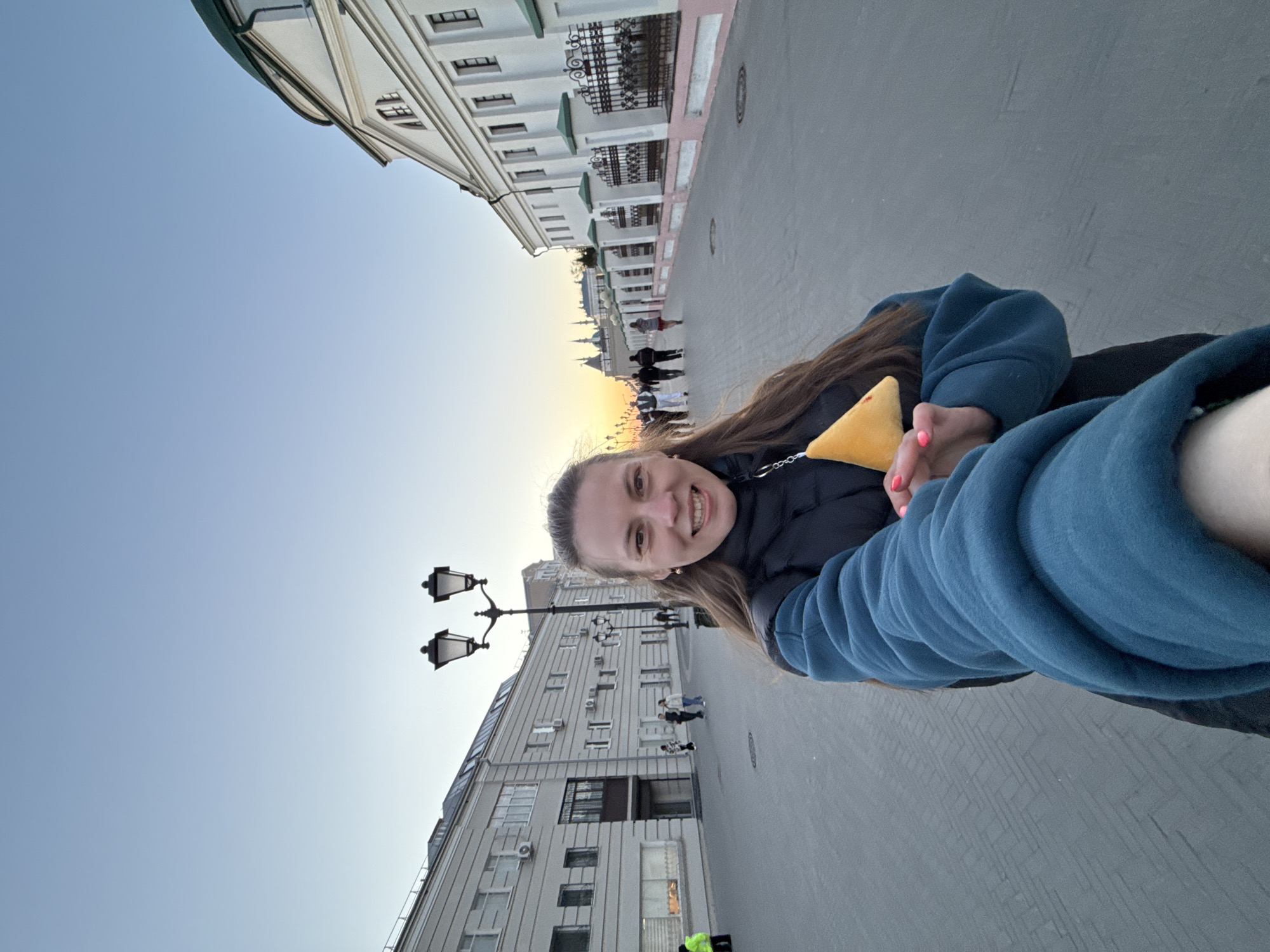 A selfie captures a woman smiling at the camera, holding a yellow, triangular object in her left hand. She is wearing a blue jacket over a black coat. The setting is a city street in Kazan, Russia, as evidenced by the distinctive architecture. In the background, a row of white buildings with green roofs and ornate black ironwork lines one side of the street, while a taller gray building with multiple windows is on the other side. Several people can be seen walking in the distance, near what appears to be a sunset on the horizon. A black lamppost stands near the woman. The ground appears to be paved with gray concrete.