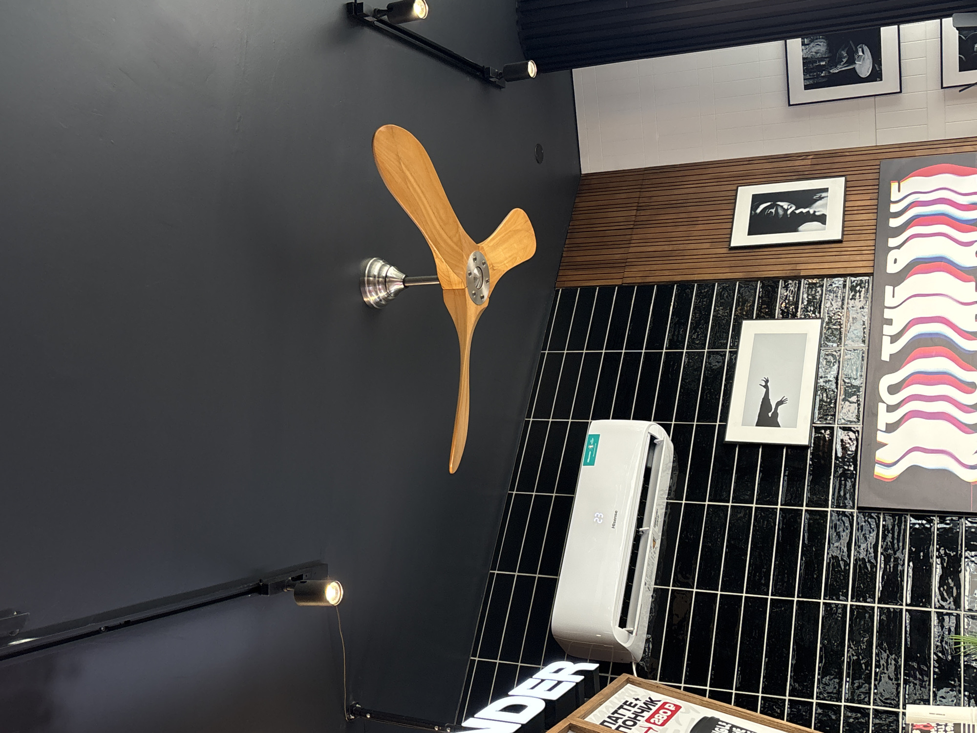The image shows an interior wall in Perm, Russia, featuring a mix of decorative elements and utilitarian fixtures. The wall is divided into a section painted black and a section covered in vertical black tile with light grout. A wooden propeller sculpture is mounted on the black painted wall. A modern air conditioning unit is installed on the tiled wall. Framed black and white photographs are displayed on the wall, alongside a poster with colorful wavy lines. Track lighting fixtures are mounted on the ceiling and wall. The image also includes a glimpse of retail signage in the lower part of the picture.