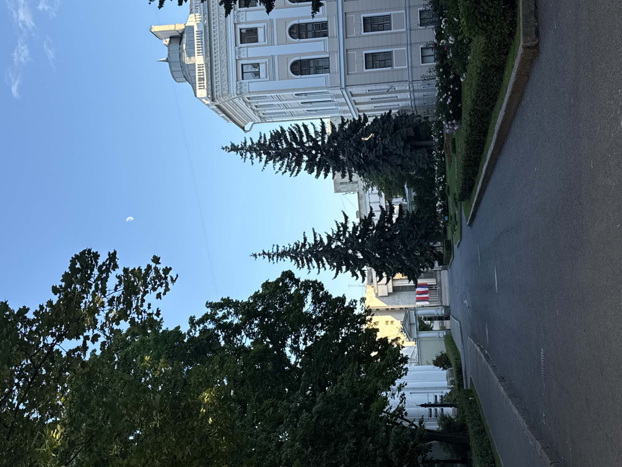 The image depicts a street scene in Saint Petersburg, Russia, with a focus on architecture and nature. A large, light-colored building with classical architectural elements, including columns and ornate detailing, dominates the upper portion of the frame. The building has multiple windows and a rounded dome-like structure on its left side. In front of the building, there is a row of mature trees, including tall evergreen conifers and deciduous trees with lush green foliage.
A paved road runs alongside the building and trees, with a manicured hedge and flowerbeds bordering the sidewalk. The road appears to be empty of vehicles and pedestrians. Two flags, red, white, and blue, are visible in the distance, attached to poles in front of another building. The sky is clear and blue, with a sliver of the moon visible on the left side of the image. The lighting suggests it is either early morning or late afternoon. The overall impression is one of a serene and grand urban landscape, highlighting the juxtaposition of historical architecture and natural greenery. There is no visible text in the image.