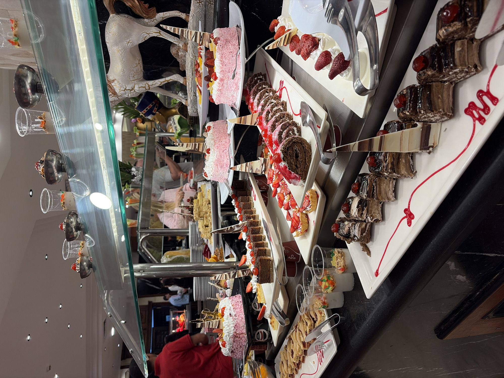 The image shows a dessert display at what appears to be a buffet, possibly in a hotel or resort. A selection of cakes, pastries, and other sweet treats is arranged on white platters and trays, some decorated with fruits like strawberries and cherries. Some individuals are visible in the background, suggesting the buffet is open and people are present. The desserts are behind a glass divider. Based on the prompt, this is likely within a location in Sharm El Sheikh, Egypt. The image captures a variety of pastries and desserts commonly found in a buffet setting.
