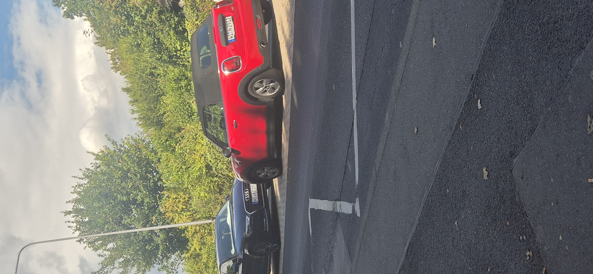 In Wiesbaden, Germany, two cars are parked on the side of a road. The car on the left is a dark-colored Audi sedan, and the car on the right is a red Mini Cooper convertible. Both cars have license plates visible. The red Mini Cooper's license plate reads "WI JN 2912", and the Audi's license plate reads "WI AZ 29". The cars are parked on a paved surface next to a tree-lined area. The sky is visible above the trees, with some clouds present. The road has white lines painted on it.