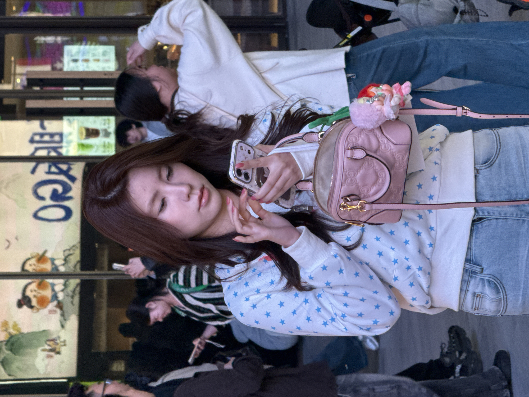 This image depicts a street scene in Liangjiang, China, focusing on a young East Asian female in the foreground. She is approximately in her mid-20s, with long, dark brown hair. She wears a white long-sleeved sweatshirt patterned with numerous light blue stars and light-wash blue jeans. Both hands hold a silver-colored smartphone, possibly an iPhone, and her right thumb is positioned over the screen. Her fingernails are painted pink. A small, light pink quilted handbag, featuring a prominent "GG" logo, is slung over her left shoulder. A pink plush doll with multi-colored hair and glitter embellishments is attached to the handbag.
In the blurred background, several other individuals are visible. To the main subject's left, a male wearing glasses is partially depicted. Further back, another East Asian female in a horizontal striped shirt is looking down, possibly at a phone. To the right, an East Asian female with her dark hair tied back in a ponytail, wearing a light-colored top and blue jeans, has her right arm raised, seemingly adjusting her hair or shading her eyes. A male with dark, curly hair is visible further back, looking towards the left. The backdrop consists of a building with large glass windows. These windows display a large cartoon graphic of two plump figures with rosy cheeks and top-knots, stylized blue Chinese characters, and illuminated advertisements showcasing beverages. The lighting indicates an evening or indoor setting with significant artificial illumination.