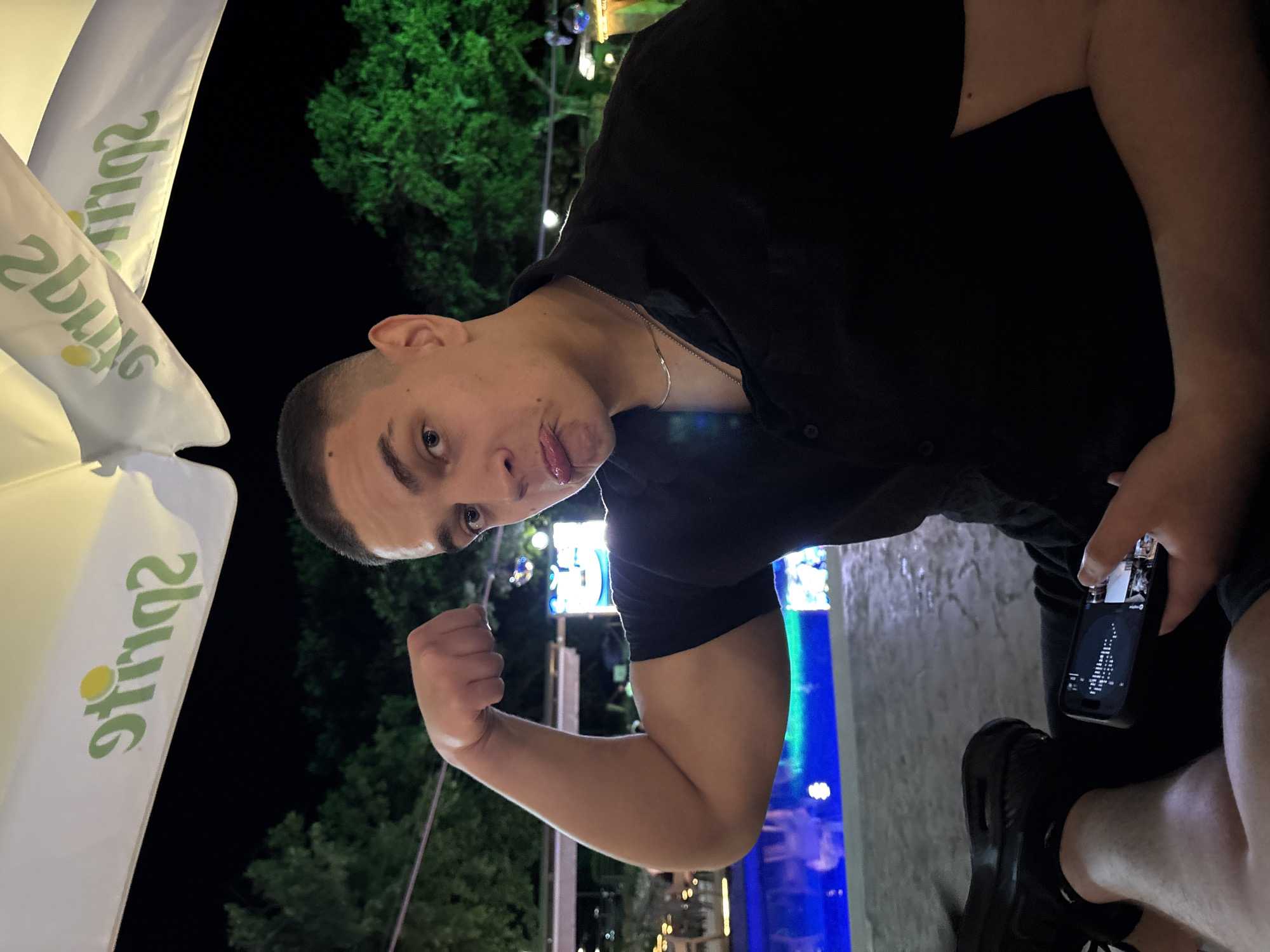 A young man with a buzz cut and a black collared shirt is posing for a photo outdoors at night. He is flexing his bicep and making a pouty face. He is wearing a silver chain necklace. In his lap, he holds a smartphone displaying images. In the background, a swimming pool with blue and green lights is visible, along with trees and what appears to be outdoor seating area under a white umbrella with the "Sprite" logo. The scene is likely in Varna, Bulgaria, given the context of the prompt.