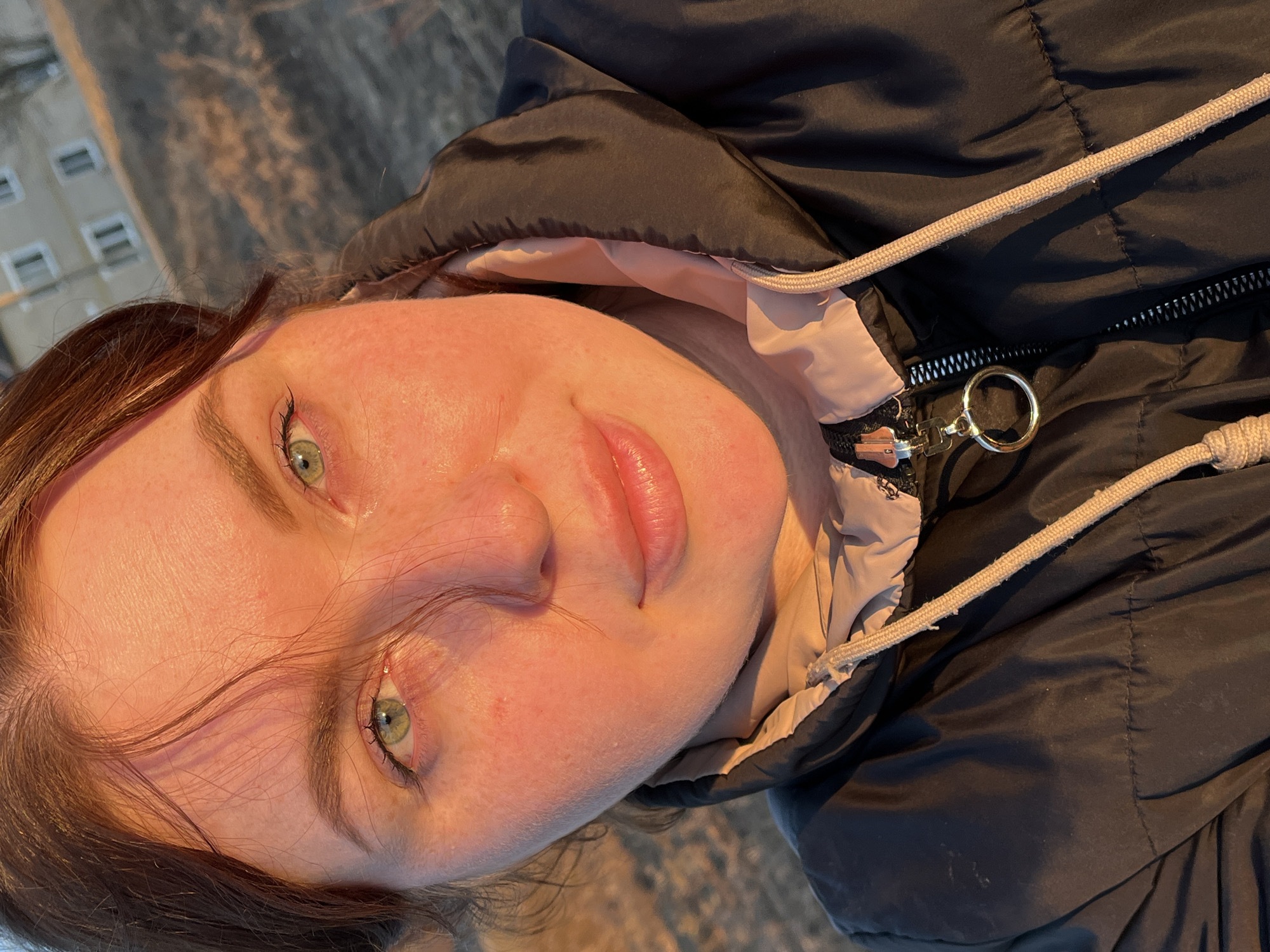 A close-up, angled view captures a young woman wearing a black jacket with a light pink lining and a drawstring hood. The zipper is partially open with a circular pull tab. She has fair skin, reddish cheeks, green eyes, and light brown hair. The background features a blurry view of a building, suggesting an outdoor setting. The location is Павловка, Russia.
