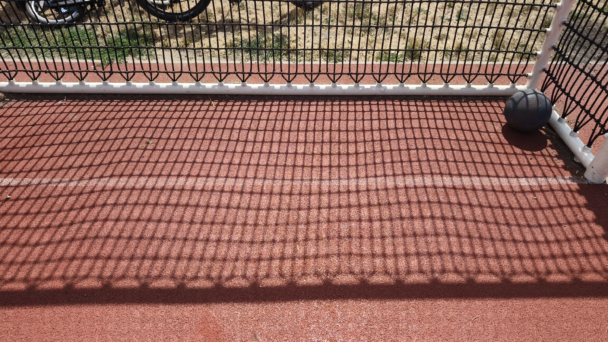 The image captures a close-up view of a red, textured outdoor sports court, likely a basketball or futsal court, with a black net casting a distinct grid shadow across the surface. The net is supported by a white horizontal bar and white vertical posts, forming a portion of a goal or barrier. A dark gray ball rests near the right edge of the frame, positioned within the shadow of the net and against the white post. In the background, out of focus, a black bicycle with visible spokes and tires is partially seen behind a section of the black net. The ground beyond the net appears to be dry grass and dirt. The overall scene suggests an outdoor recreational area. The location is identified as Vienna, Austria.