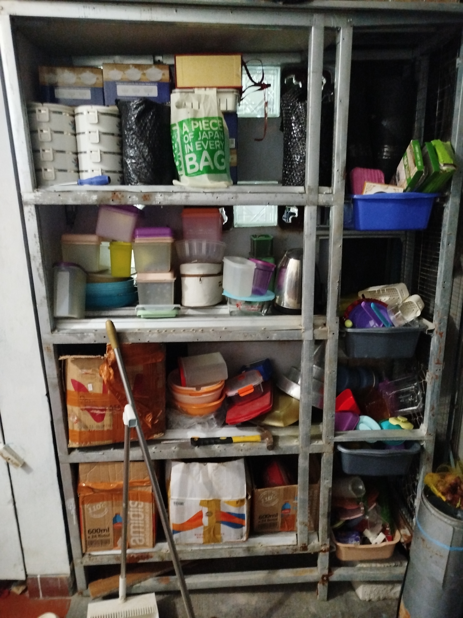 This is a photograph of an industrial metal shelf unit filled with various household items, plastic containers, boxes, and kitchenware. The shelf unit appears to be old and somewhat rusted, with multiple levels containing an assortment of objects. On the top shelf, there are several boxes and a green plastic bag. The second shelf from the top holds several white cylindrical containers, a black plastic bag, and a green plastic bag with text. Below that, there are various colored plastic containers, bowls, and a stainless steel kettle. Further down, more boxes, bowls, and plastic containers are visible. A broom is standing upright in front of the shelf unit. The overall environment appears to be a storage area or a workshop, possibly in a residential or commercial setting. The image does not contain any specific elements that would indicate a location in Bandungrejo, Indonesia, nor does it depict any people or specific actions.