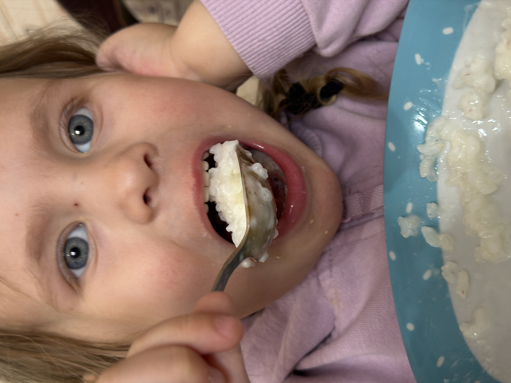 This close-up, eye-level image, captured indoors in Chaikovsky, Russia, features a young child, likely a girl, engaged in eating. The photograph is rotated, presenting the child's head at a significant tilt. She has light-colored hair and striking blue eyes, which are open and looking towards the viewer. Her fair skin is visible, and she is dressed in a light purple or lilac long-sleeved top.
The central action shows a metallic spoon, laden with white, creamy food resembling rice porridge, partially inserted into her open mouth. Her lips are a natural pink hue. To the child's right, a light blue bowl with white polka dots is visible, holding additional portions of the same porridge. The background is softly blurred, indicating an indoor setting, likely a home. The child appears focused on her meal. There is no visible text in the image. The specific time of day is not discernible, but the scene clearly depicts a mealtime.