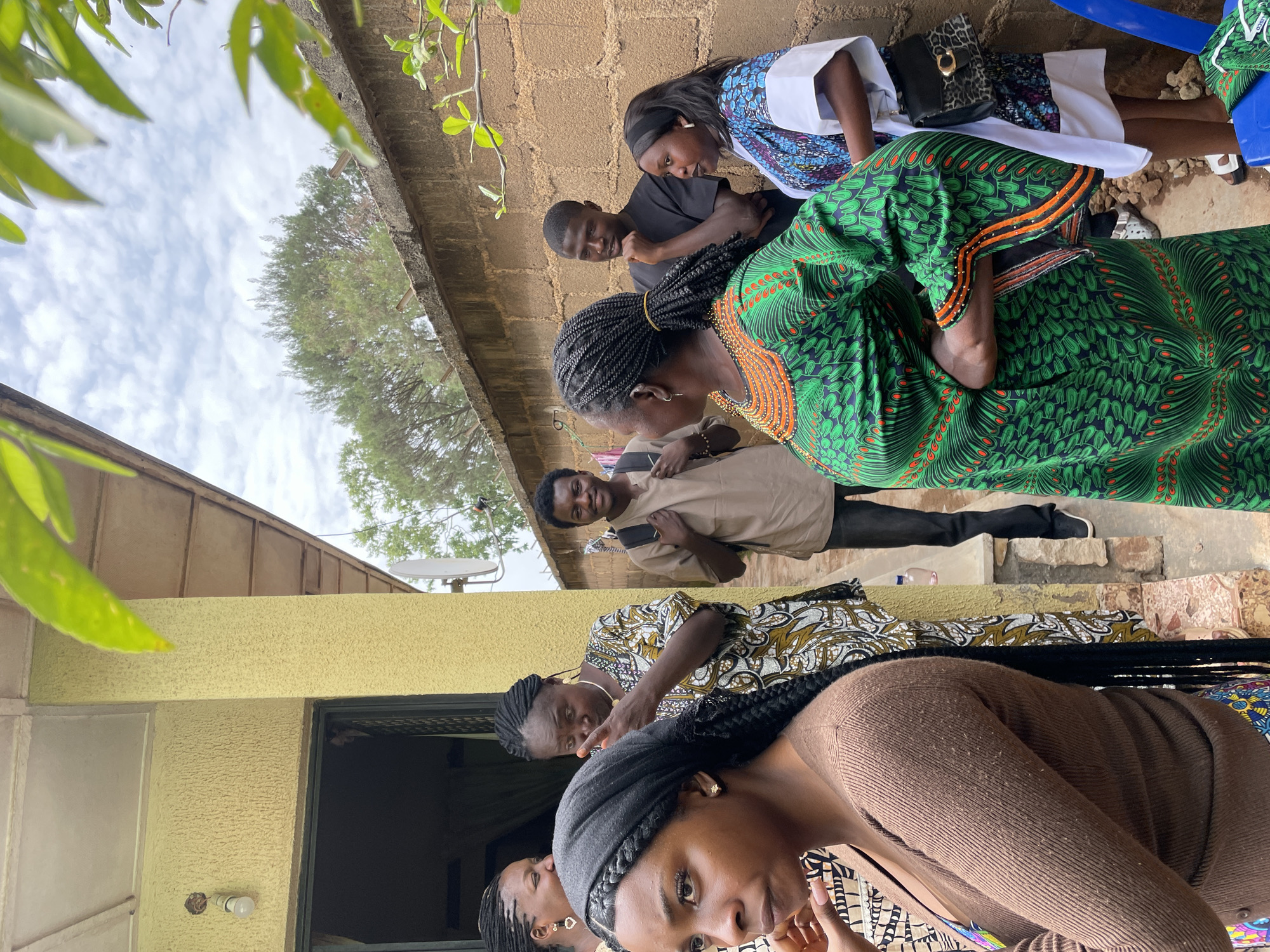 The photograph shows a group of people outdoors, likely in Jos, Nigeria, due to the style of clothing and the architectural features visible. The scene is captured from a slightly low angle, with individuals standing and interacting. Several people are wearing traditional African attire with vibrant patterns and colors. Some appear to be looking down or engaged in conversation. The background includes a brick wall, a building with a yellow exterior, and some foliage visible in the upper portions of the frame. A satellite dish is attached to the building. The ground appears to be a paved or stone surface. A blue plastic chair is also partially visible.