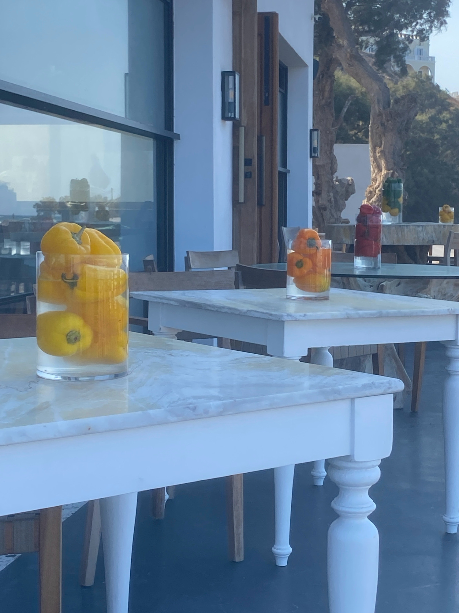 The image depicts an outdoor seating area with several tables, likely at a restaurant or cafe, in Thira Municipal Unit, Greece. In the foreground, a white marble-topped table with a white base holds a clear glass vase filled with bright yellow peppers submerged in water. Another white table with a similar marble top is visible behind it. In the mid-ground, a darker-colored table supports a tall red vase containing what appears to be red peppers or tomatoes. To the right of this, another clear glass vase holds yellow peppers and possibly limes. Further in the background, more tables and chairs are present, as well as large glass windows reflecting the environment. Beyond the seating area, a large, gnarled tree stands prominently, and in the distance, an architectural structure with arches can be seen. The overall setting suggests a bright, sunny day.