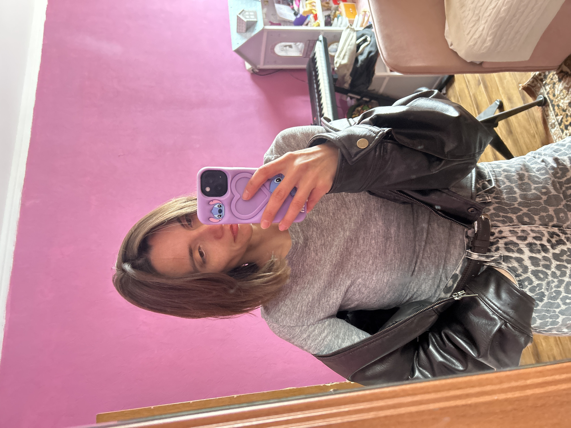 A young woman is taking a selfie in front of a mirror. She is wearing a gray long-sleeved shirt and a black leather jacket. The case of her phone is purple and has a cartoon character on it. The background includes a pink wall, a white dollhouse, a keyboard, and a chair. The location is not identifiable as Moscow, Russia.