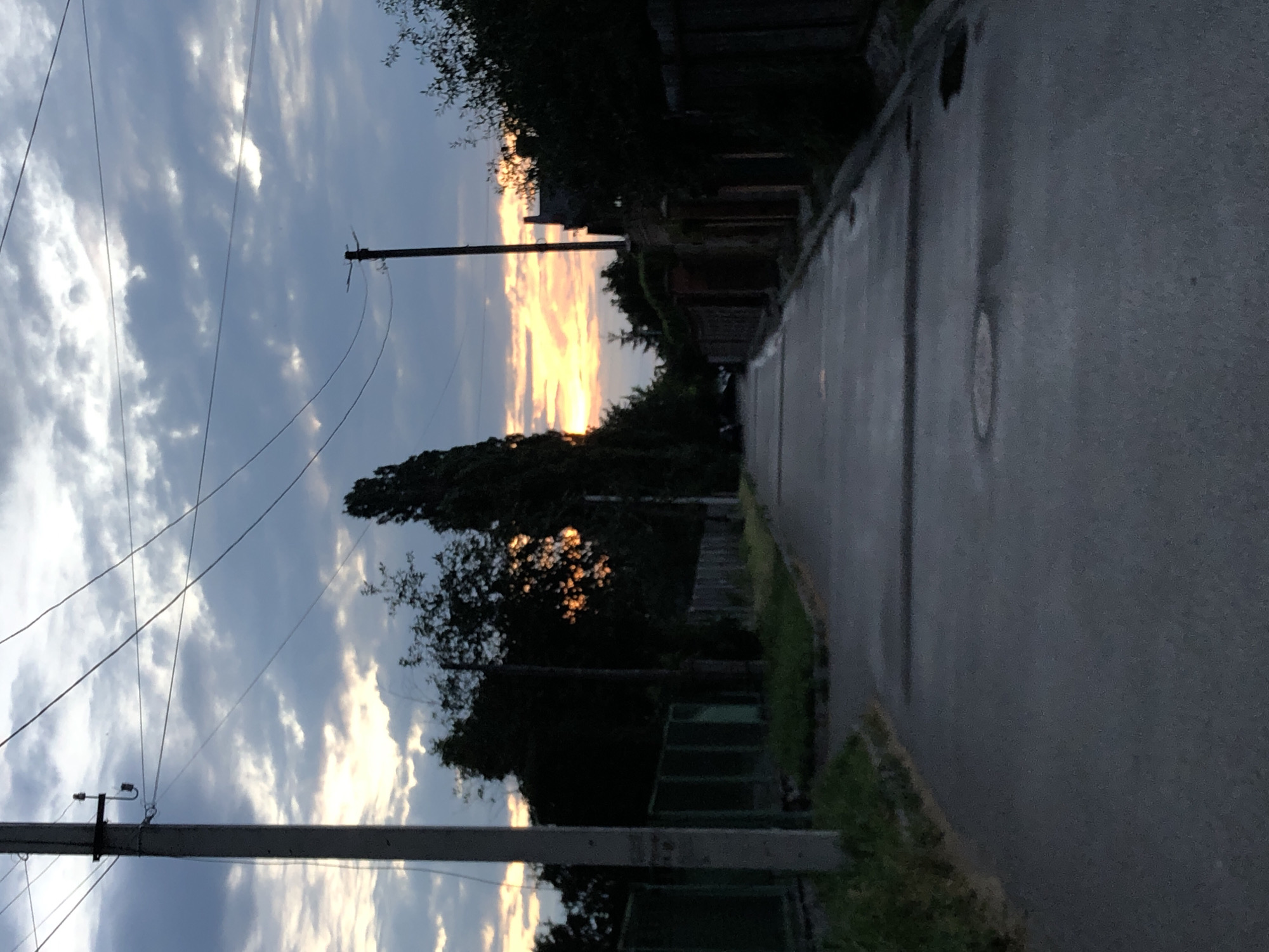 This is a vertically oriented, slightly blurry, low-angle, outdoor photograph of a street scene during twilight. The sky dominates the left half of the frame, filled with a dramatic mix of dark grey and light blue clouds, interspersed with streaks of vibrant orange and yellow light from a setting or rising sun, suggesting either dawn or dusk. Power lines stretch across the sky, supported by at least two utility poles visible in the frame.
The right side of the image depicts a paved street that recedes into the distance. The pavement appears wet, reflecting the dim light and suggesting recent rain or dew. Along the right edge of the street, there is a grassy verge with some low-lying vegetation. Further back, a series of low fences and shadowed structures, possibly houses or outbuildings, are visible, partially obscured by dark silhouettes of trees and foliage. The overall mood is somewhat melancholic and serene, characteristic of the transition between day and night. There is no visible text in the image.
