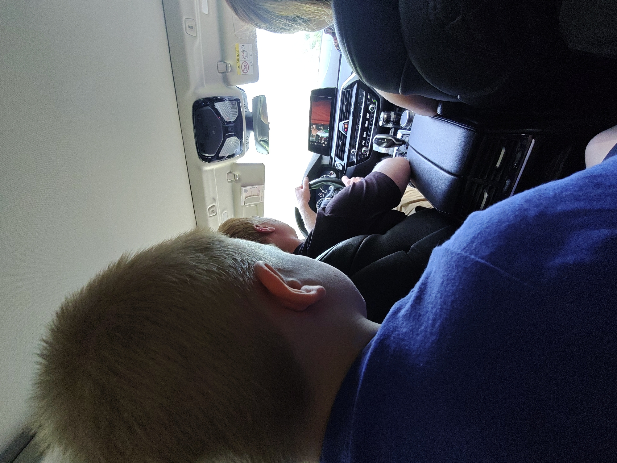 A group of people are inside a car on a road, possibly on a trip. The image is taken from the perspective of someone in the backseat. A young boy with blonde hair and wearing a blue shirt is in the foreground, his head and shoulder visible. To his right, a man with brown hair is driving, and a woman with blonde hair is in the passenger seat. The car's interior features a digital dashboard with a screen displaying a video, air conditioning vents, and a gear stick. The car appears to be moving through a rural or suburban landscape, possibly near Kurakino, Russia, with the bright daylight suggesting it's daytime.