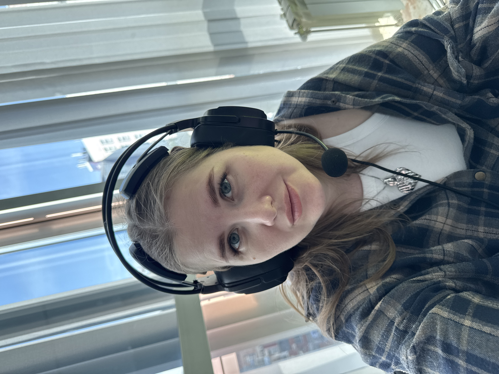 A young woman with light-colored hair and blue eyes is the central subject of the image, captured in a close-up, slightly upward-angled selfie that is rotated 90 degrees clockwise. She wears a black over-ear headset with a boom microphone positioned near her mouth. Her attire includes a white t-shirt under an open plaid shirt featuring a blue-grey and tan pattern. A metallic pendant, possibly a whistle or a detailed charm, hangs on a necklace around her neck.
She gazes directly at the camera with a calm and pleasant expression, marked by a slight, soft smile. The background reveals an indoor setting, illuminated by bright daylight coming through a window covered by horizontal blinds. Beyond the window, a clear blue sky and faint outlines of distant buildings are visible, indicating it is daytime with good weather. While no specific landmarks are discernible, the location context is Perm, Russia. The headset suggests she may be engaged in activities like remote work, online communication, or gaming. There is no visible text in the image.