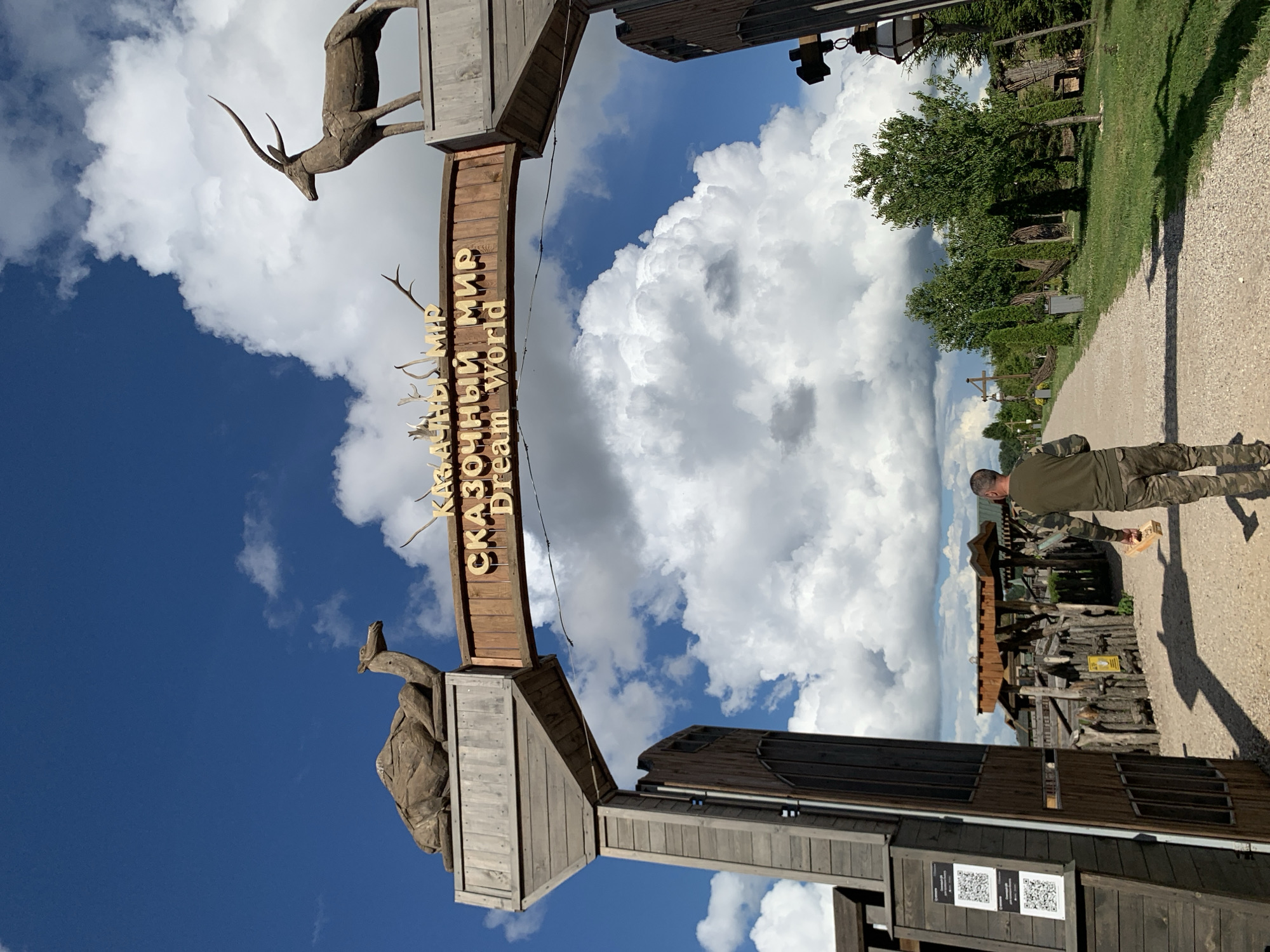 The image captures the entrance to "Dream World," a themed park, likely in Малахавецкі сельскі Савет, Belarus, judging by the text on the sign. The entrance is an archway constructed of weathered wood. Atop the left pillar of the archway, a wooden sculpture of a camel is visible, and atop the right pillar, a sculpture of an antelope or gazelle. The park's name is displayed prominently on a wooden sign above the arch, with text in Russian "СКАЗОЧНЫЙ МИР" and in English "Dream World." A man wearing camouflage pants and a green long-sleeved shirt is walking away from the camera down a gravel path leading into the park. He is carrying a small wooden box. The background reveals more of the park's rustic architecture, including wooden fences, trees, and structures, all set against a bright blue sky with dramatic white clouds. On the left side of the entrance, there are two QR codes displayed on a wooden panel. The overall scene suggests a sunny day at an outdoor entertainment venue.