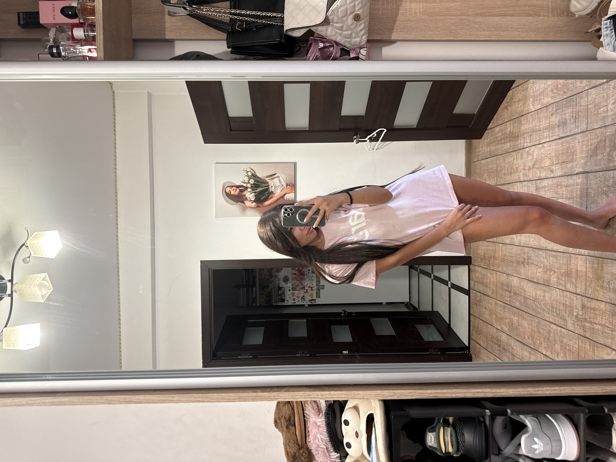A young woman with long dark hair is taking a mirror selfie in what appears to be a bedroom or dressing area. She is wearing a light pink t-shirt and no bottoms. Her left hand is holding her phone, which is positioned to capture her reflection. To her left, a mirror reflects a closet area with shelves of shoes, plush toys, and a dark brown door. To her right, another reflection shows a wooden closet with hanging bags and a shelf with perfume bottles. In the background, a framed picture of a woman holding tulips hangs on the wall. The room has wooden flooring and white walls.
