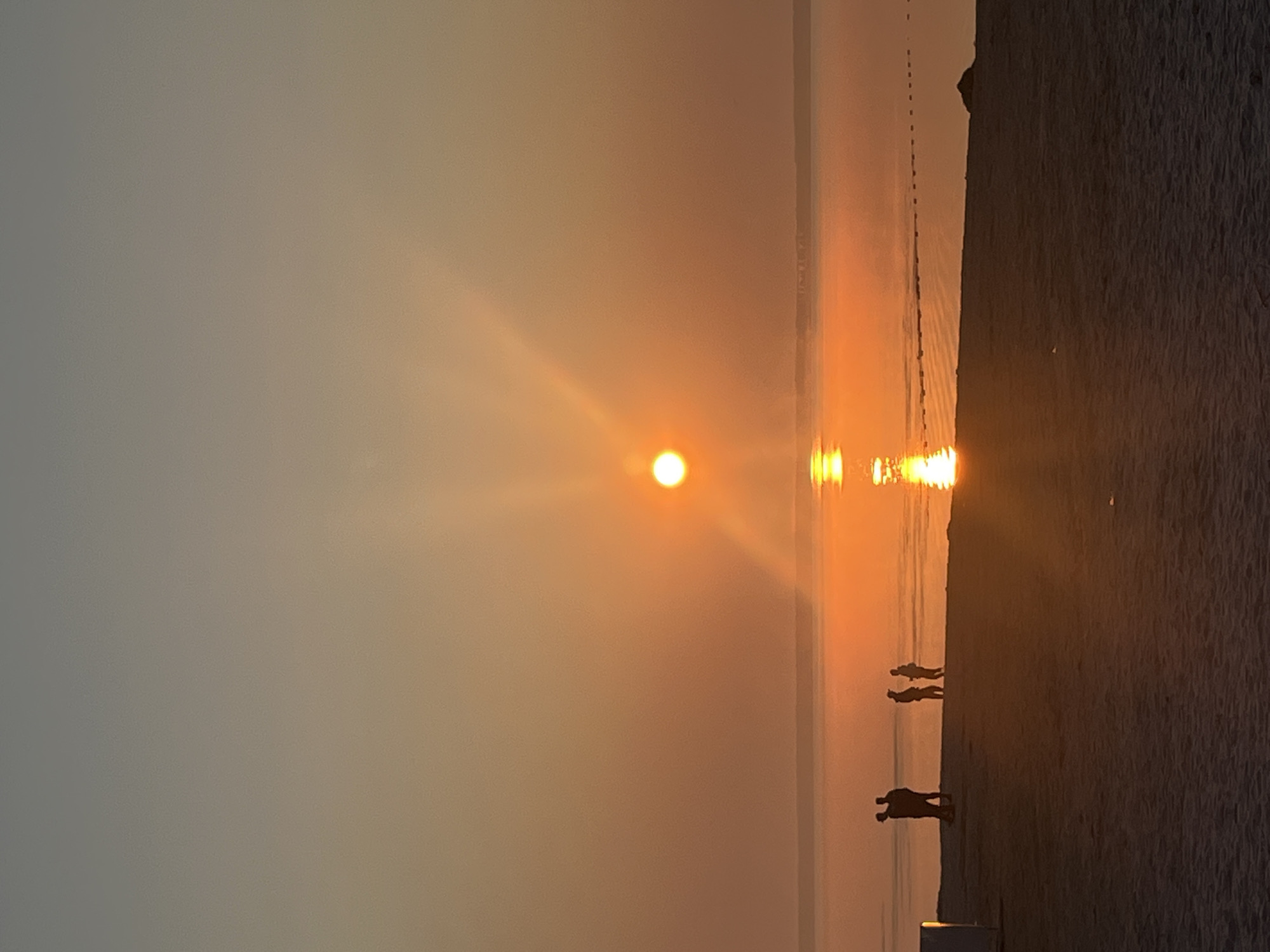 The image depicts a sunset over a body of water, likely the Gulf of Finland, from the beach in Surakovo, Russia. The sun is a bright orb low on the horizon, casting a warm orange glow that reflects on the calm water. Silhouettes of three individuals are visible on the sandy beach, facing the water and the sunset. One pair of figures stands close together on the left, while a third figure stands slightly apart to the right. The beach itself appears to be composed of sand and some scattered debris. The sky transitions from a pale orange near the horizon to a muted grey at the top of the frame.