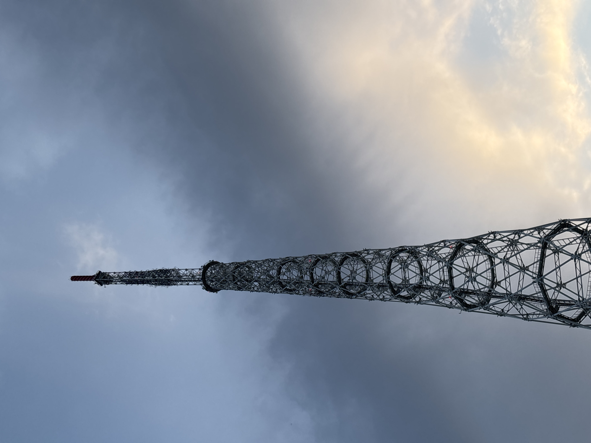 The image shows a steel lattice tower extending into a cloudy sky. The tower is composed of repeating cylindrical sections, interconnected by a complex framework. The sky is a mix of grey and light yellow, suggesting either an overcast day or twilight. The location is likely Perm, Russia, given the tower's resemblance to the Shukhov Tower on the Kama River. There are no people visible.